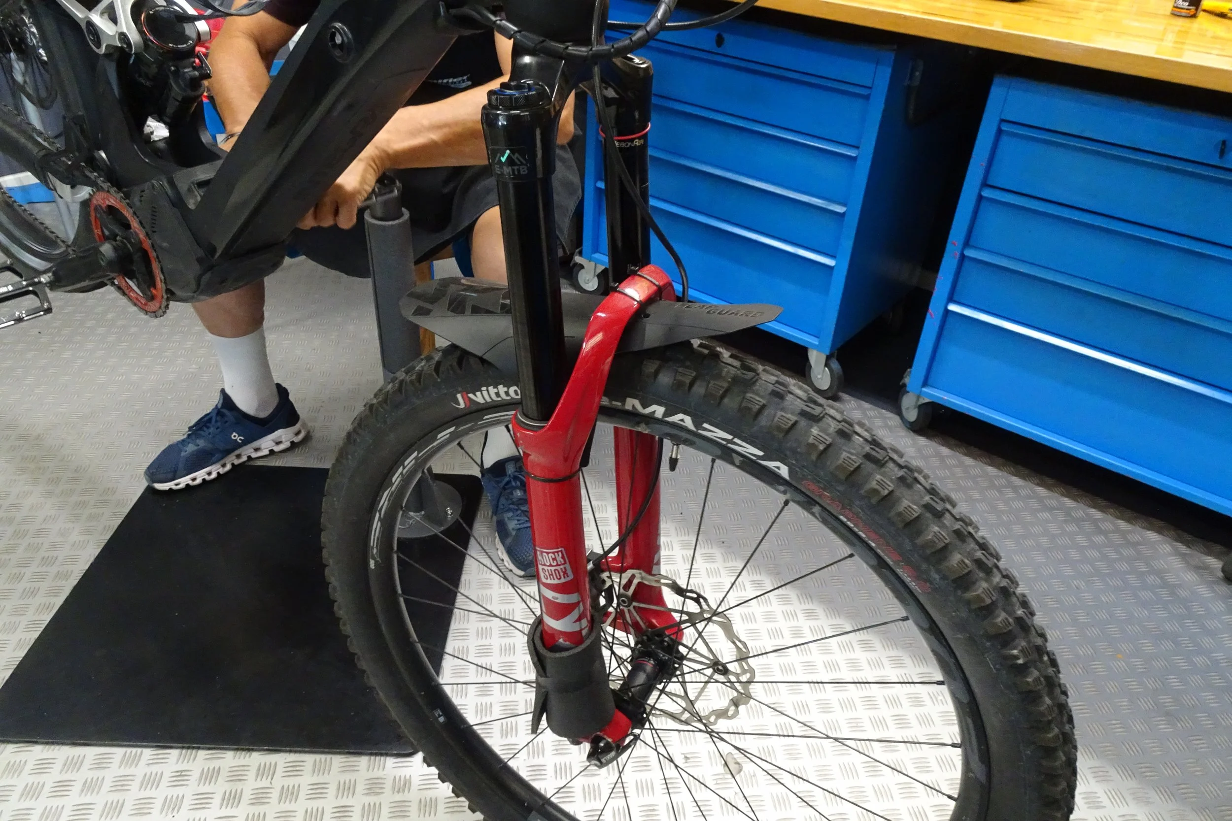 Una bicicletta da montagna rossa in un'officina, con una persona al lavoro sulla sospensione anteriore.