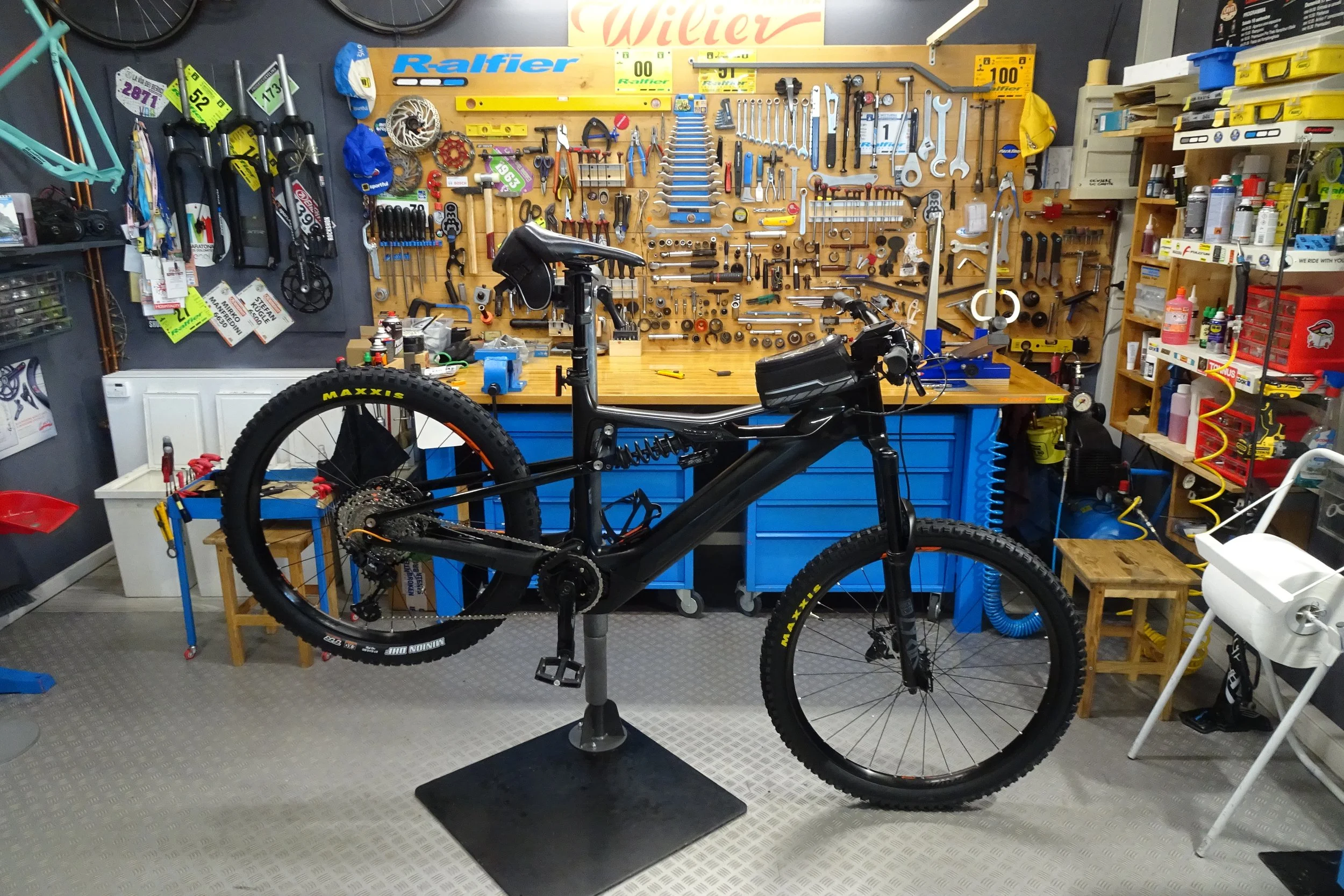 Un garage o officina con una bicicletta montata su un support. Dietro, su una parete, vari attrezzi e parti di biciclette organizzati e appesi. Sul lato destro, scaffali con varie scatole e lubrificanti.