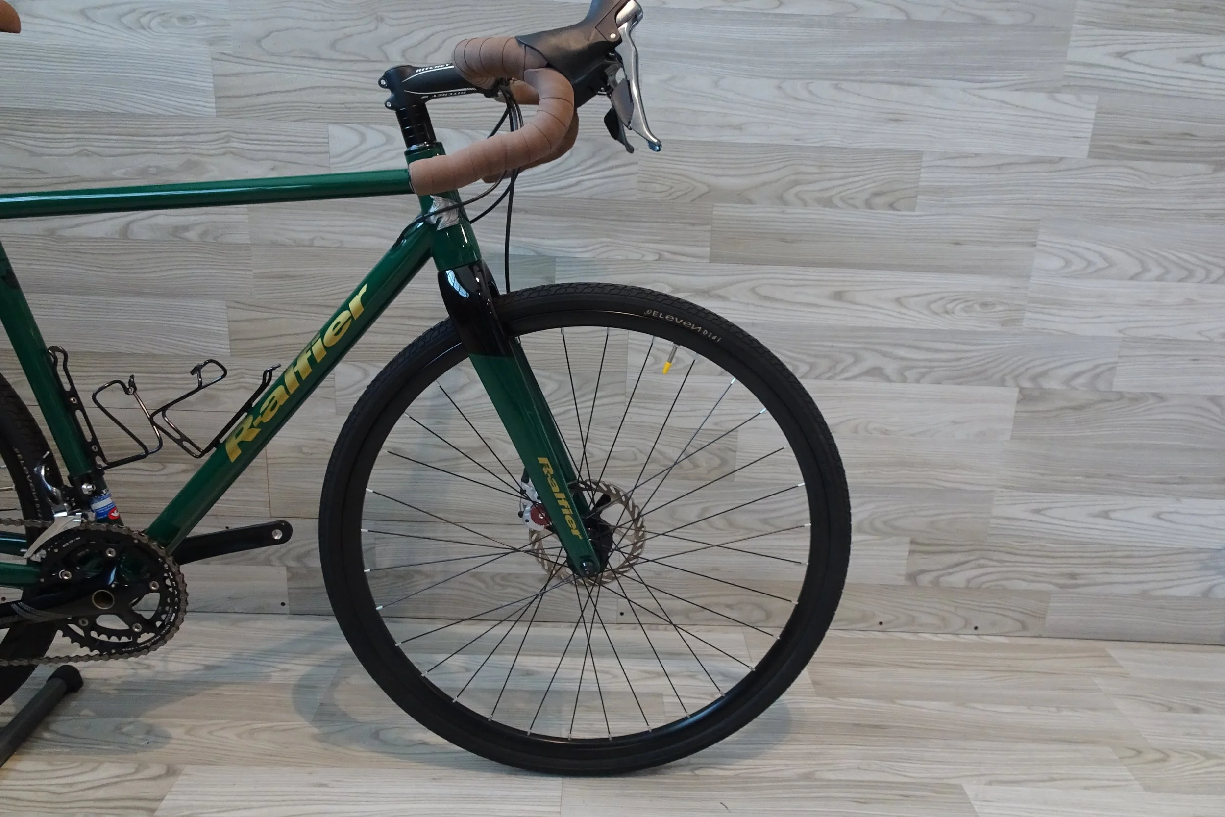 Bici da corsa Verde con manubrio in pelle marrone e freno a disco, in posizione verticale contro un muro con pannelli di legno chiaro.