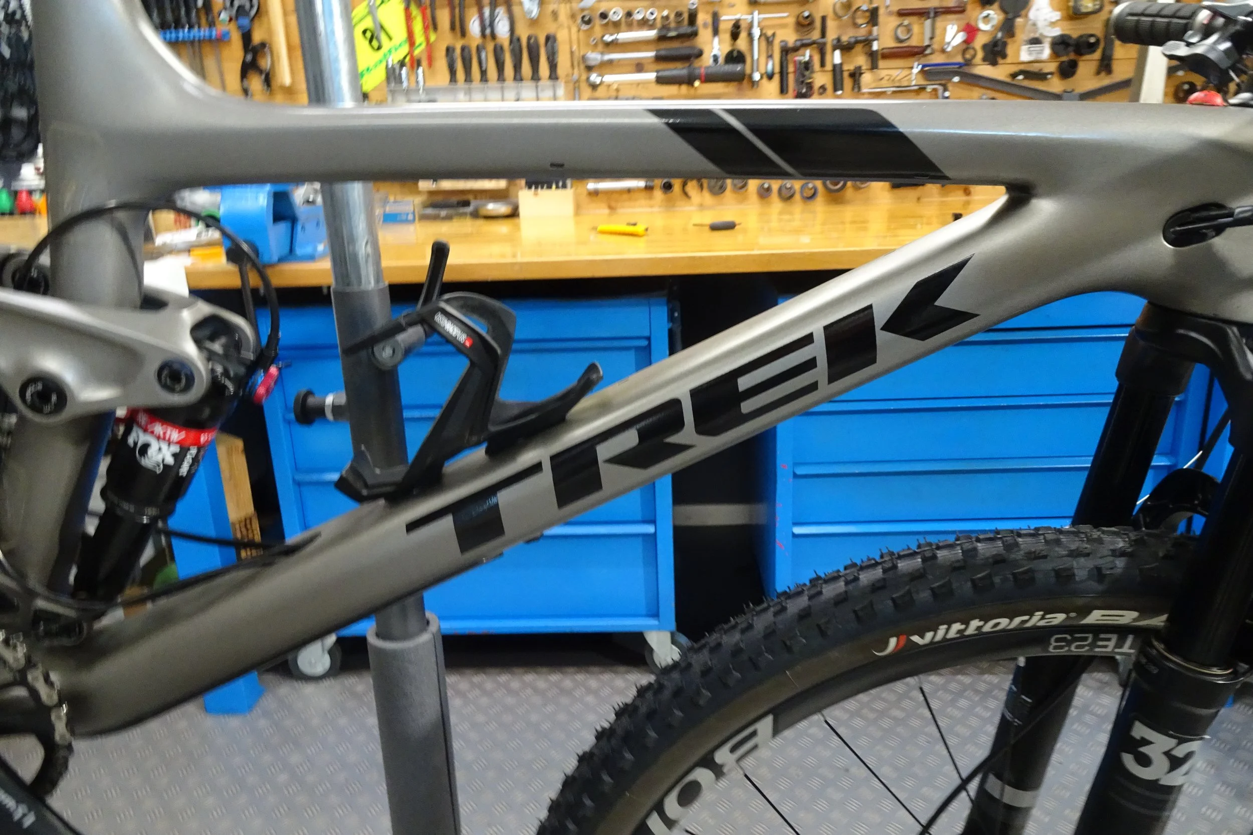 Immagine di un telaio di bicicletta di marca Trek in fase di assemblaggio in un’officina, con attrezzi appesi a una parete in background e un banco da lavoro.