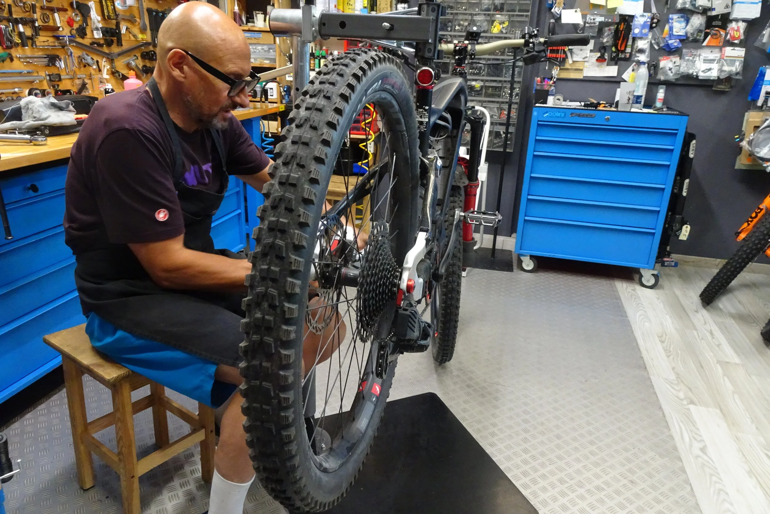 Un uomo con occhiali da vista sta riparando una bicicletta nella sua officina, circondato da attrezzi e strumenti appesi alle pareti e su un banco.