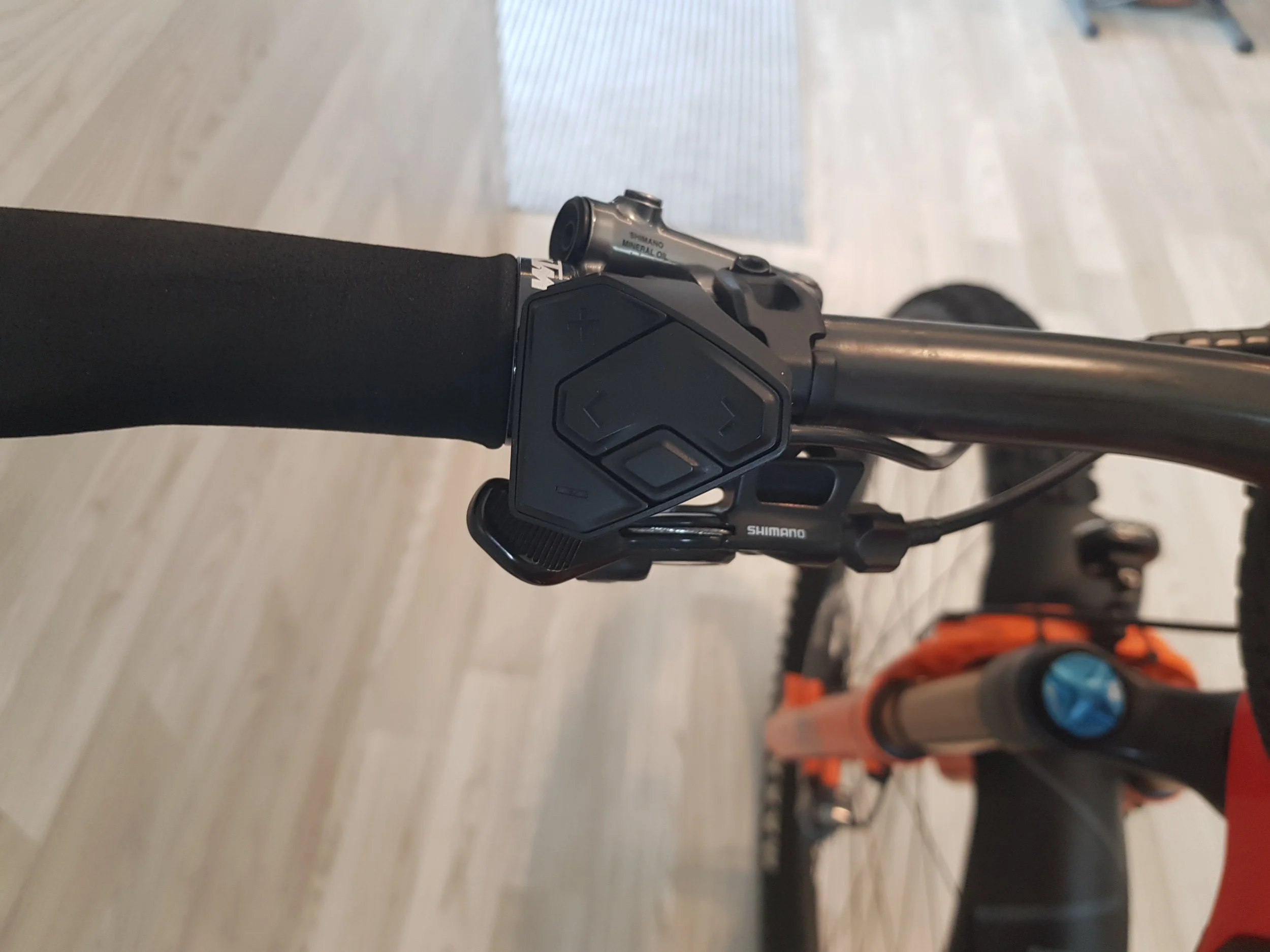 Pezzo di manubrio di una bicicletta con controllo digitale e componente Shimano, visibile da sopra, con fondo di pavimento in legno