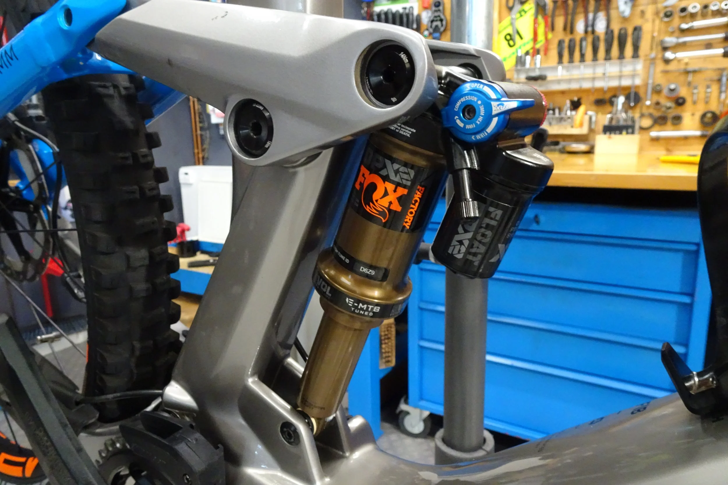 Particolare forcella anteriore di una mountain bike con sospensione a raggi X, in un workshop con strumenti da officina sullo sfondo.
