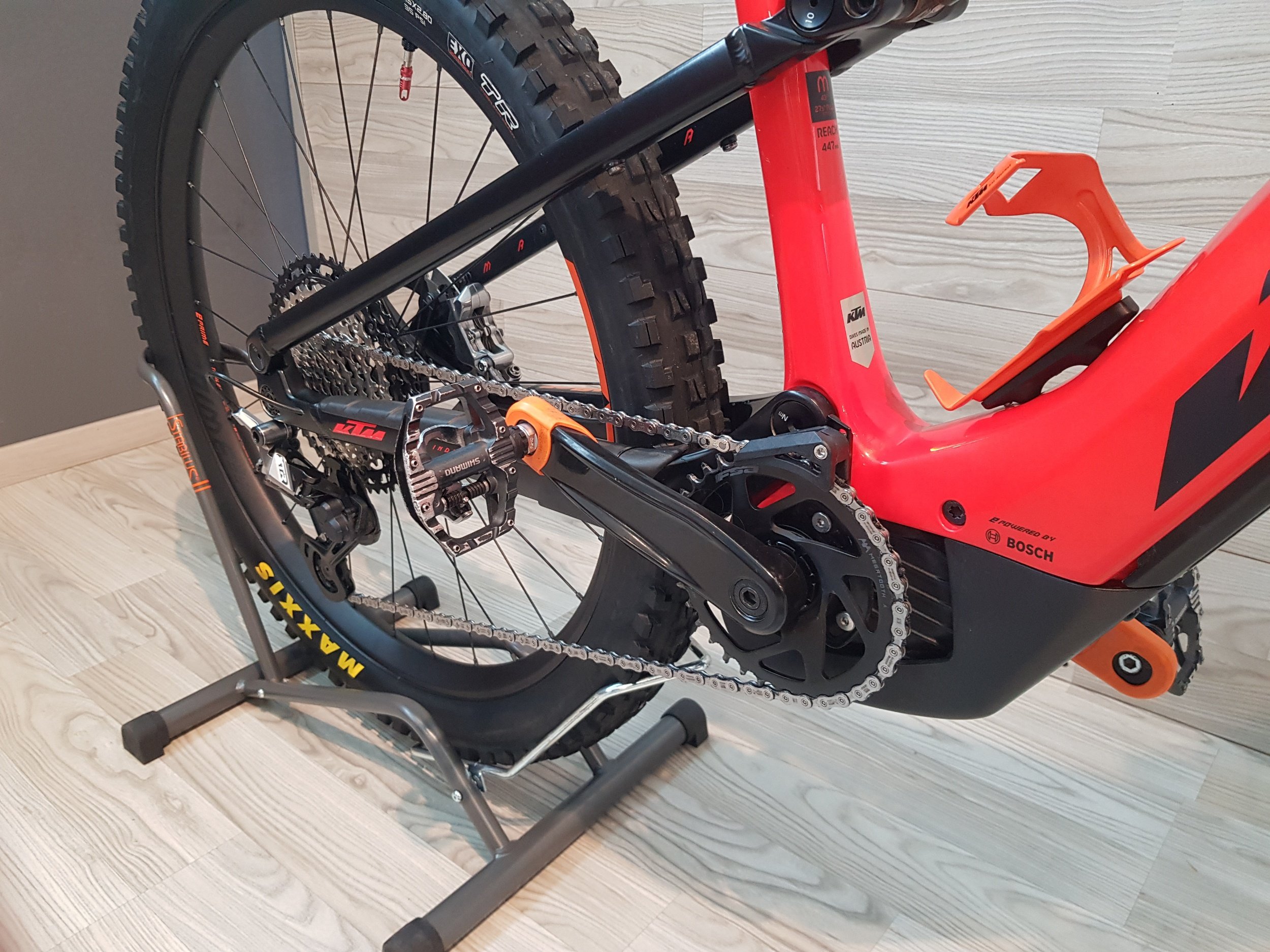 Bicicletta da enduro color rosso e nero montata su un supporto. La bici ha pneumatici grandi e tassellati, cambio e catena visibili, e pedali piatti. La bici si trova su un pavimento in legno e uno sfondo grigio.