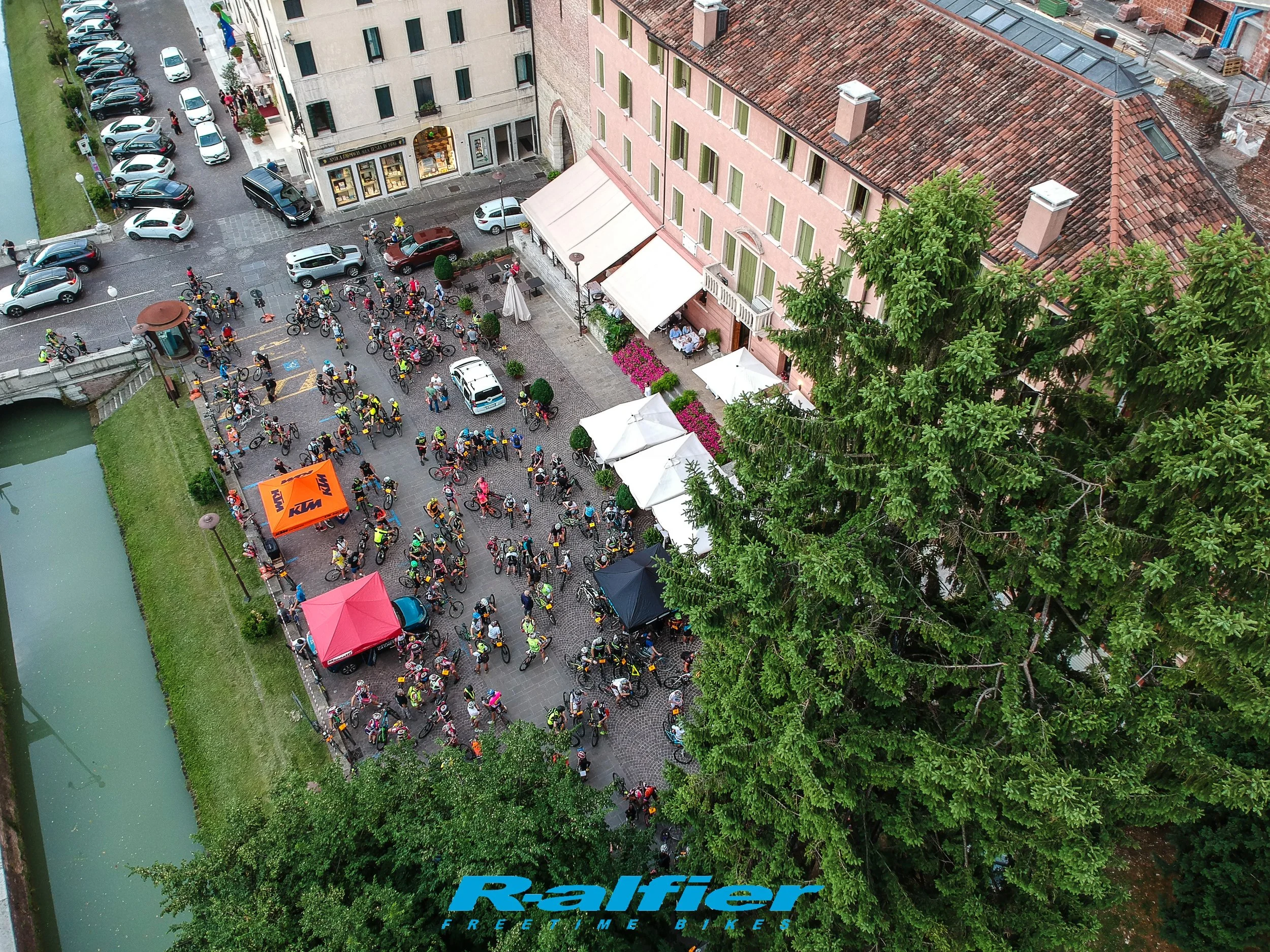 Gruppo di ciclisti riuniti in una piazza in Italia, con edifici storici sullo sfondo e alcuni negozi, alcuni tavoli con ombrelloni e grande albero verde.