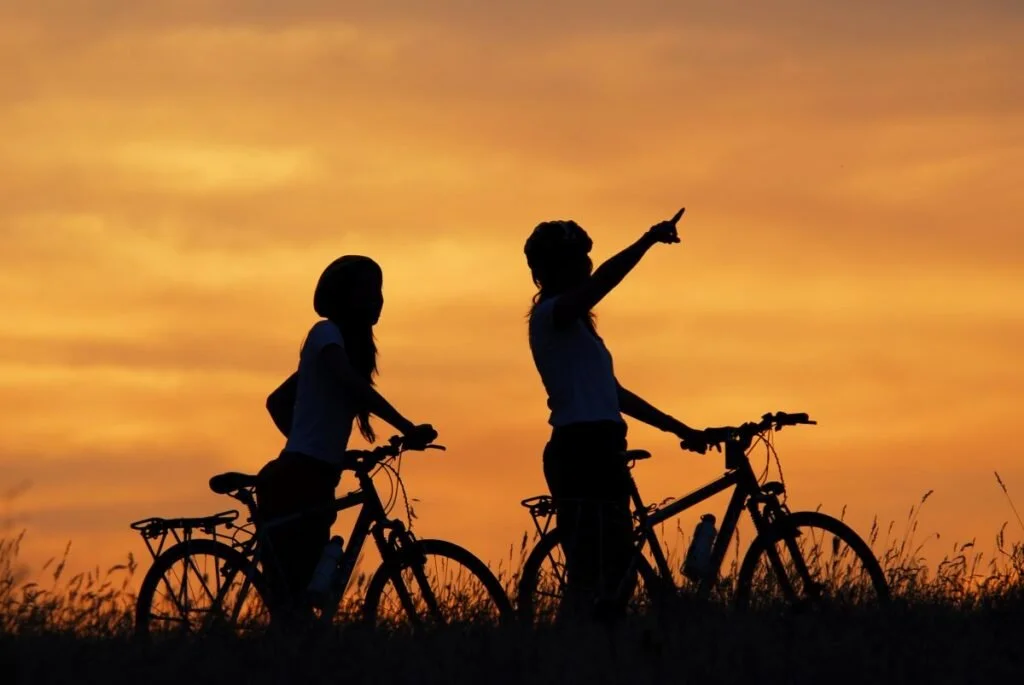 Due persone con biciclette in piedi all'aperto al tramonto o alba, silhoulette contro un cielo arancione.