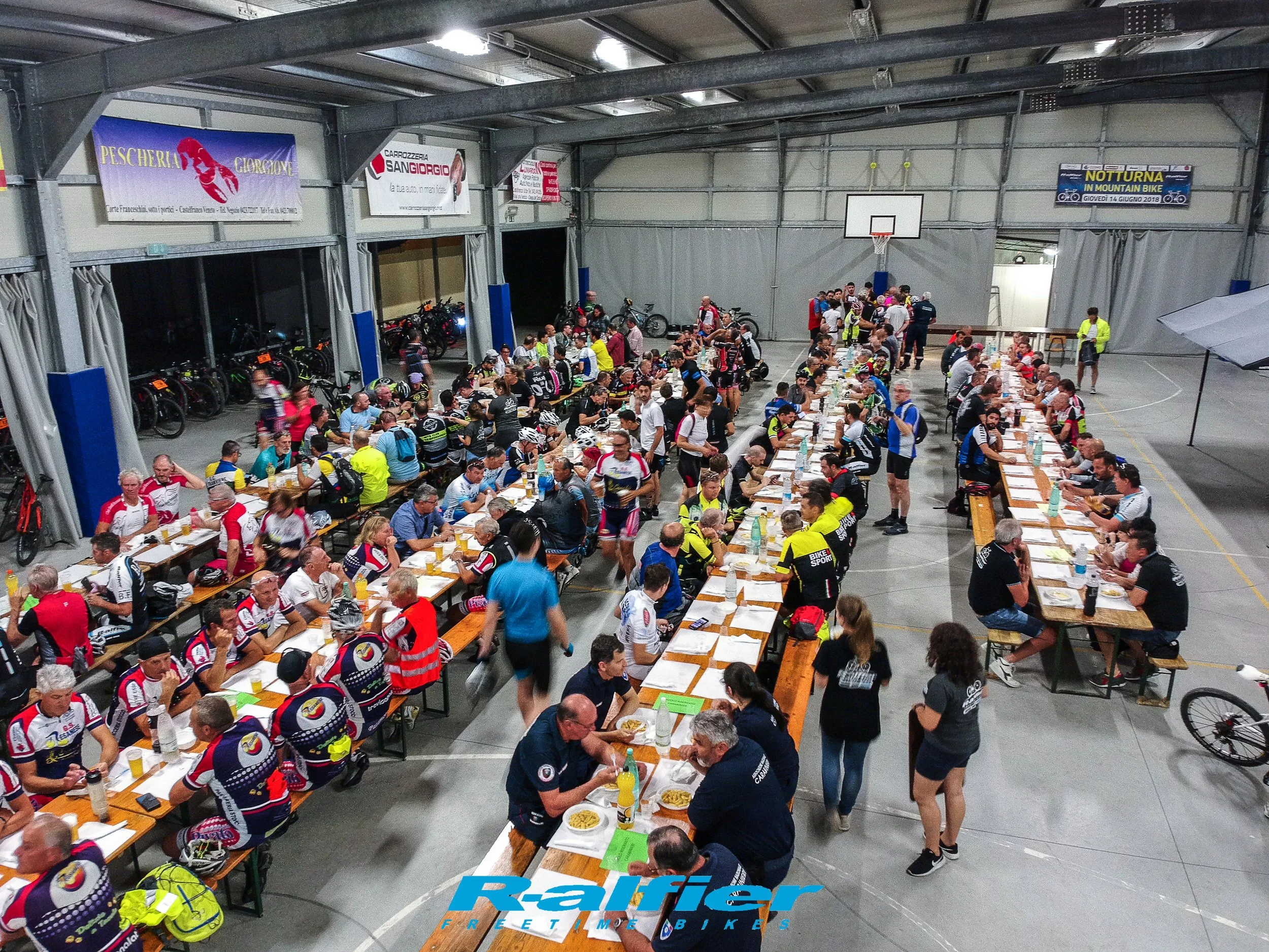 Un grande ritrovo di ciclisti e appassionati di mountain bike seduti a tavoli all'interno di una palestra o uno spazio coperto, con alcune bici appoggiate ai lati e striscioni pubblicitari sospesi sopra. Attività sociale, evento di ciclismo.