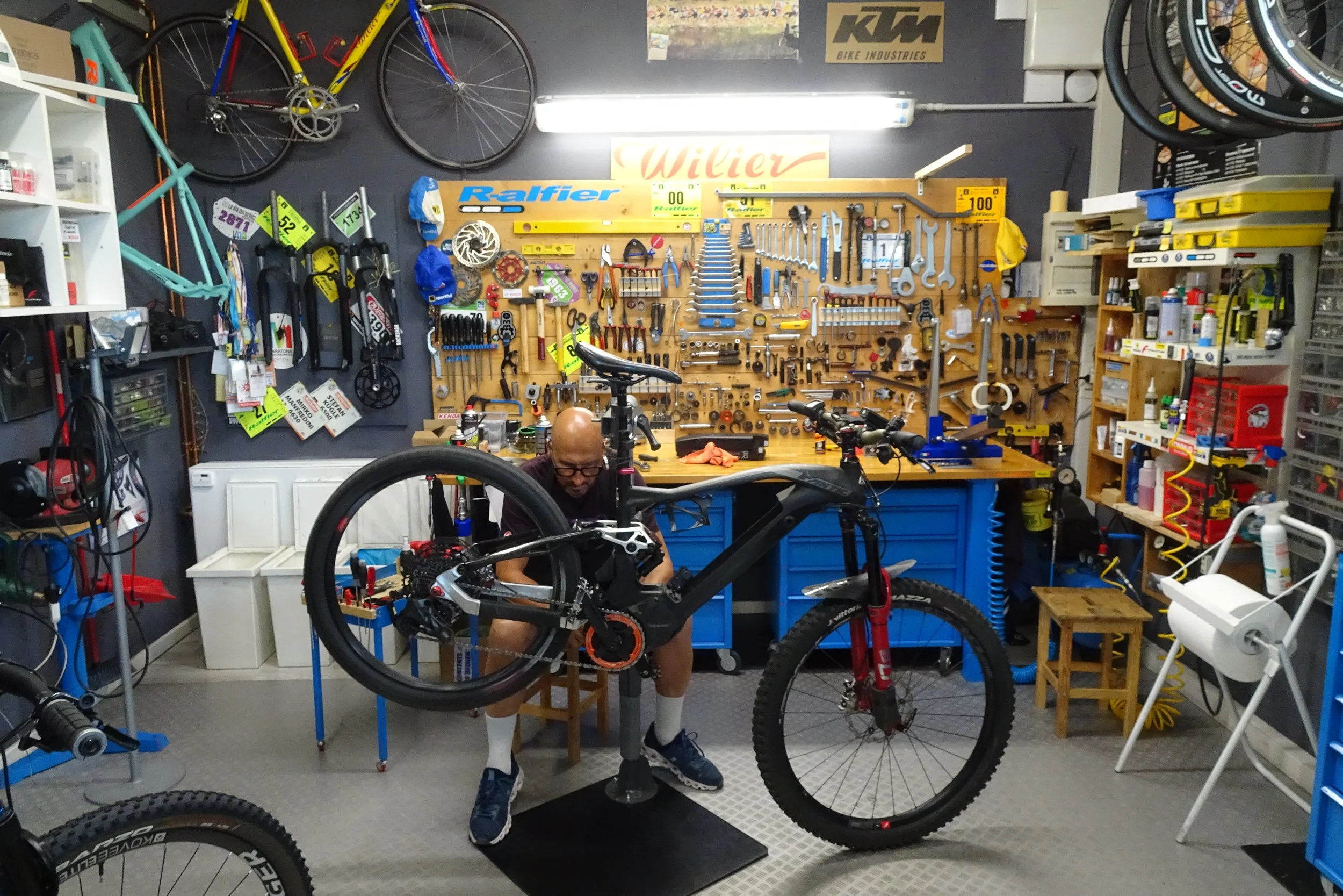 Un uomo sta riparando una bicicletta in un garage attrezzeria con diversi attrezzi e componenti appesi alle pareti.