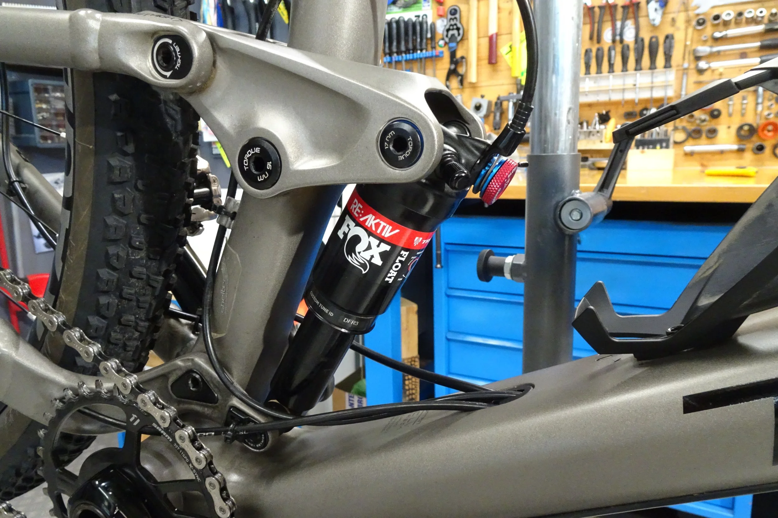 Particolare di una bici da montagna con ammortizzatore a sospensione posteriore e catena, in un'officina con attrezzi appesi sul muro sullo sfondo.