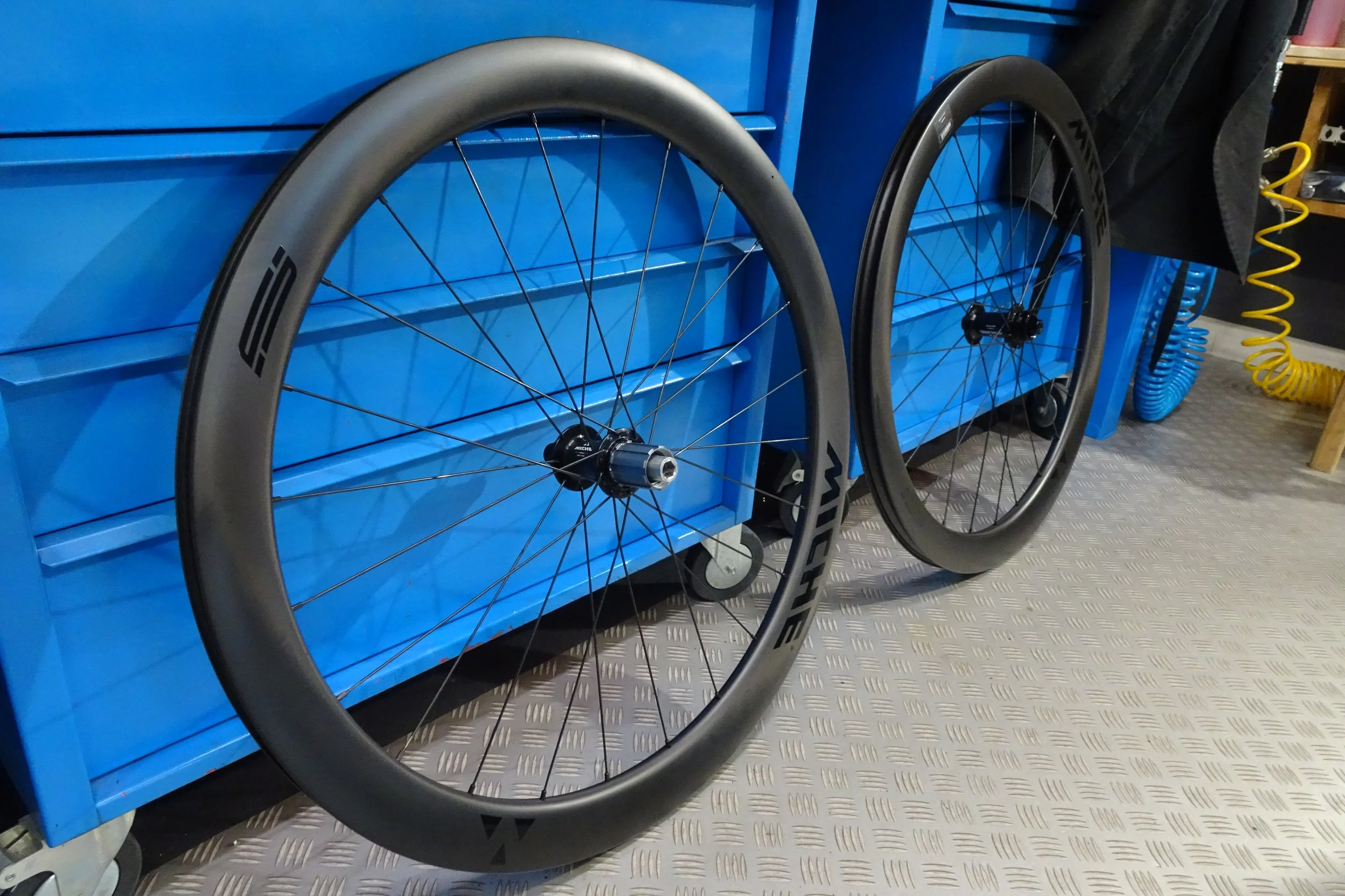 Due ruote di bicicletta nere e grigia appoggiate su una superficie metallizzata, con una mobiletto blu sullo sfondo e alcuni oggetti vari come una sedia e tubi di aerazione gialli.