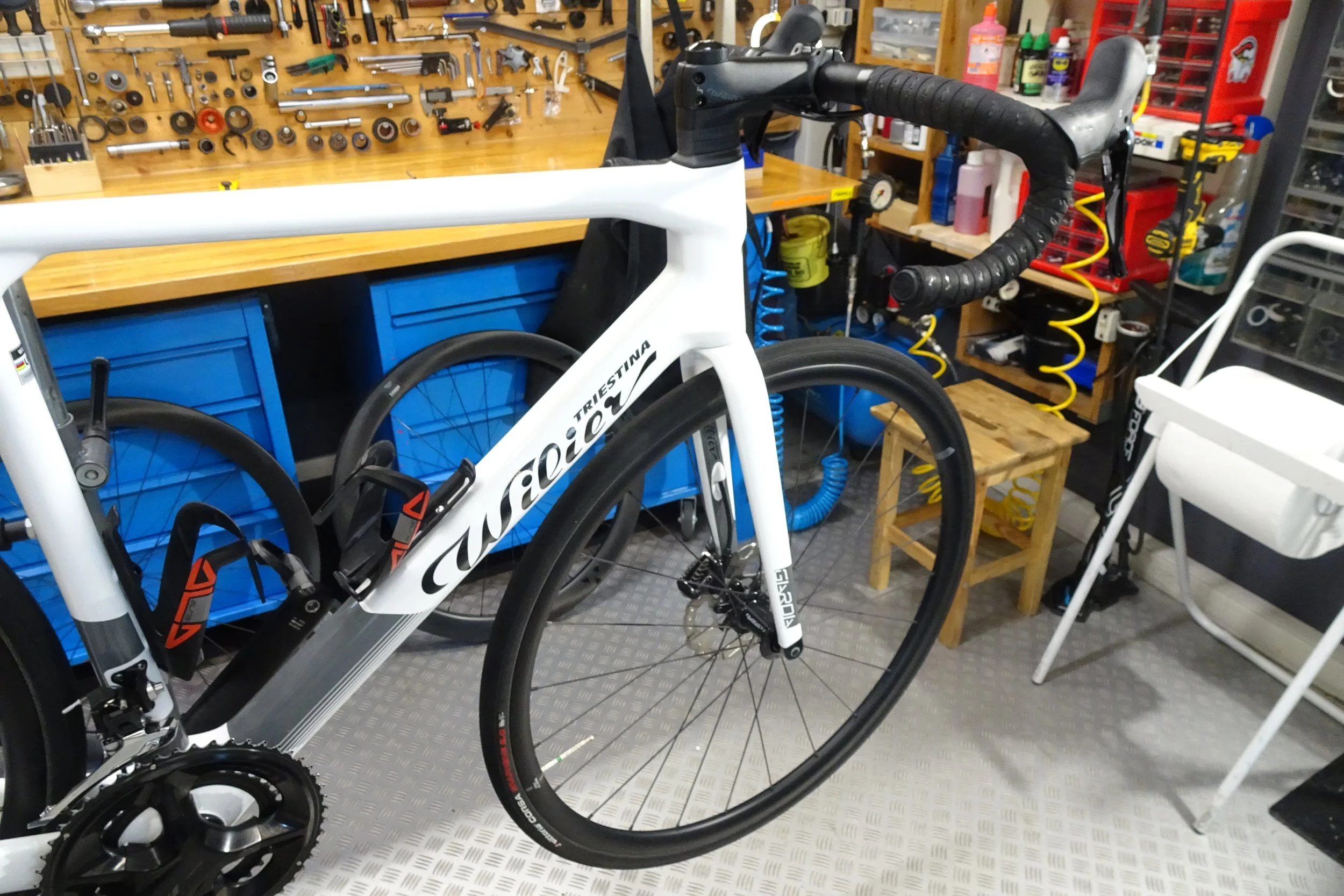 Bici da corsa bianca con dettagli neri, posizionata in un'officina con parete attrezzata con attrezzi e scaffali.