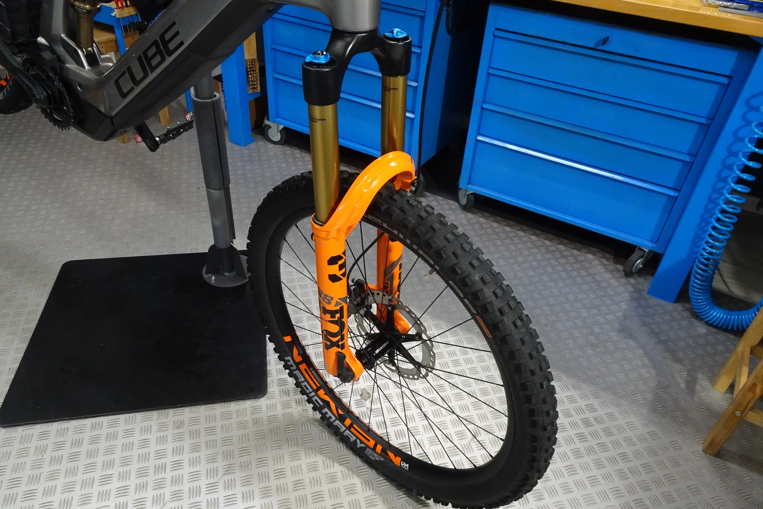 Bicicletta da montagna con forcella sospesa arancione, postata in un'officina con strumenti blu e banco di lavoro.