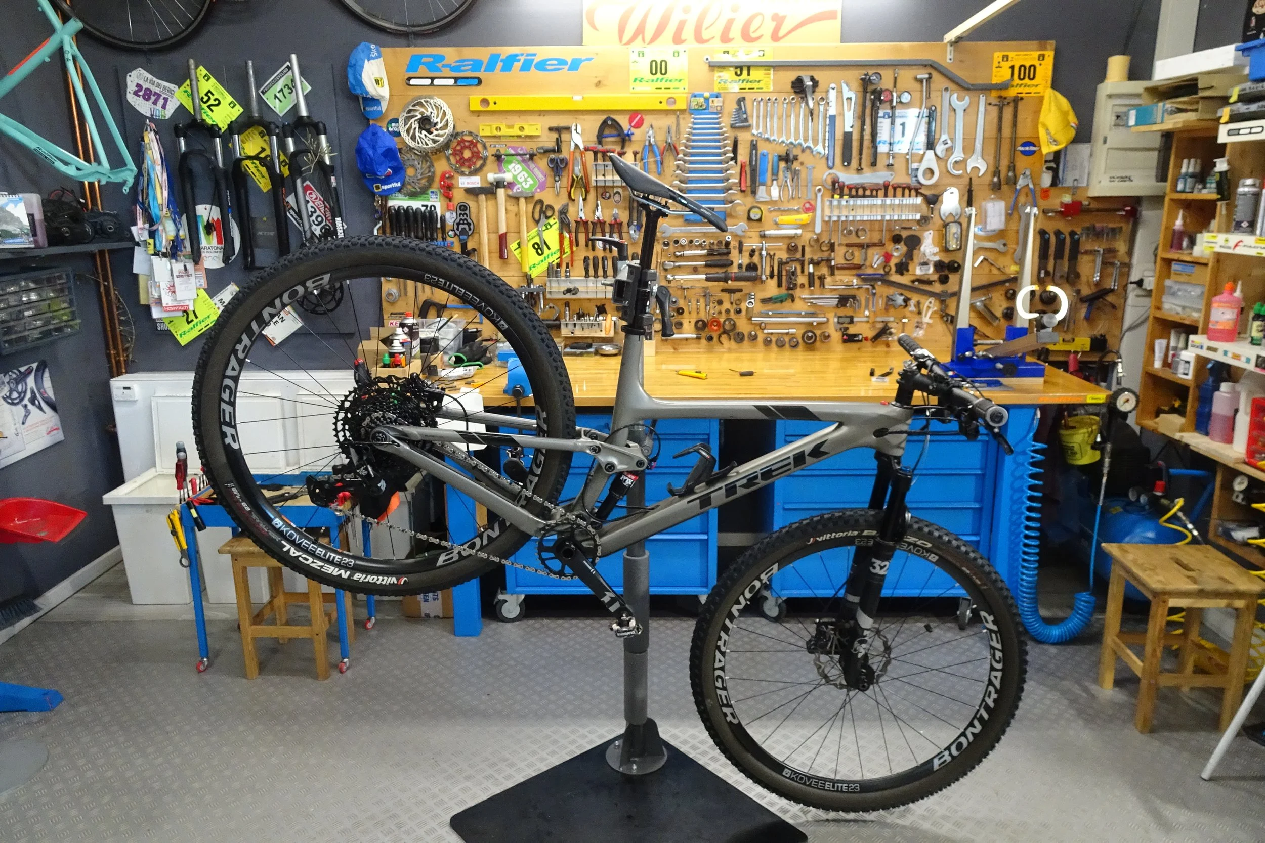 Una stanza di un'officina con un bicicletta da montagna sospesa sul support. Sullo sfondo, una parete di pannelli di legno piena di utensili e strumenti da ferramenta, tra cui chiavi, cacciaviti, e altri attrezzi. Sulla parete superiore si vede anche un'area con etichette colorate e numeri.
