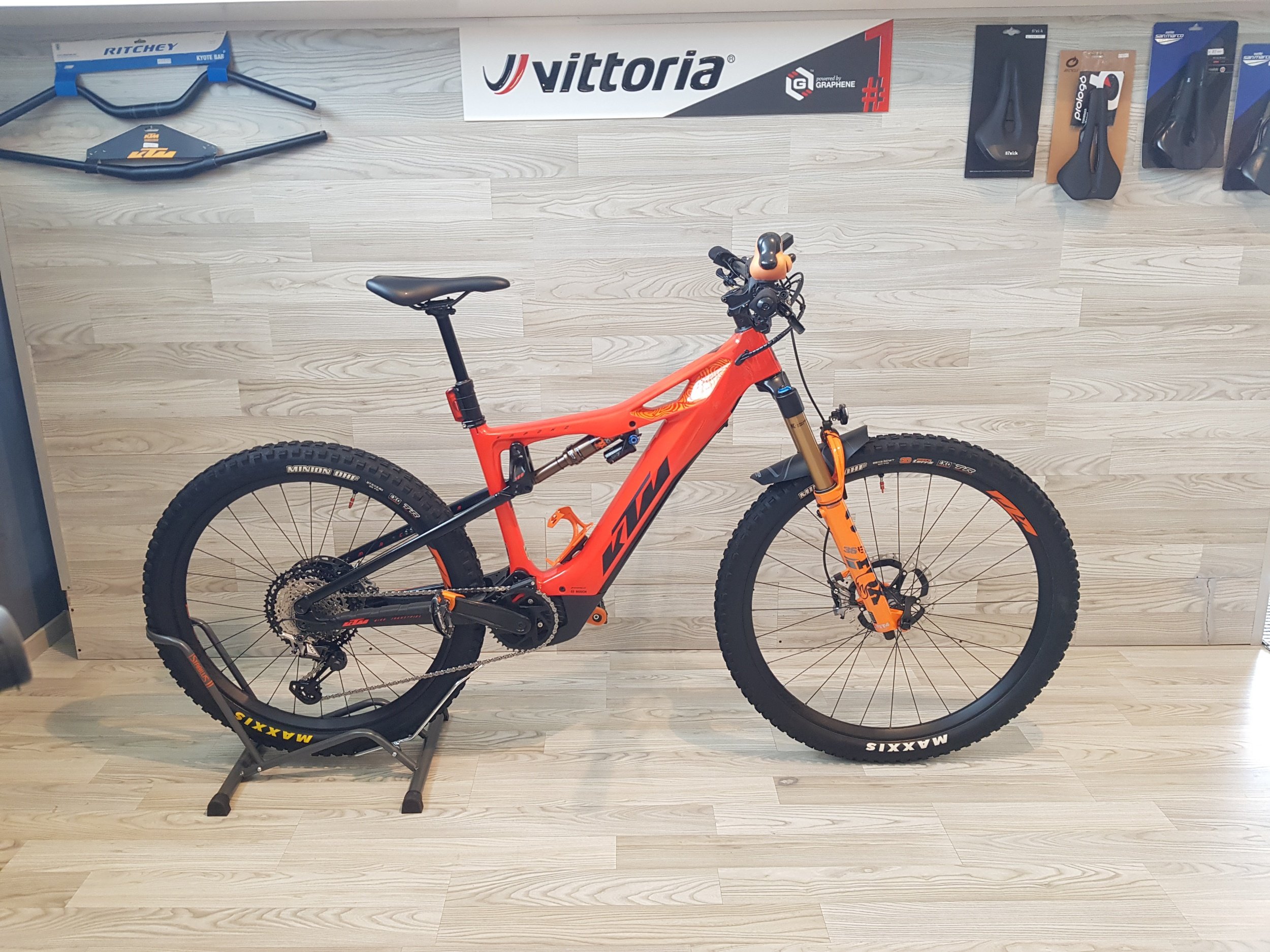 Bicicletta rossa da mountain bike su una rampa di allenamento, con accessori esposti sul muro dietro, tra cui un manubrio, un sellino, guanti e altri oggetti sportivi. Sono presenti anche pannelli pubblicitari e un banner con il logo 'Vittoria'.