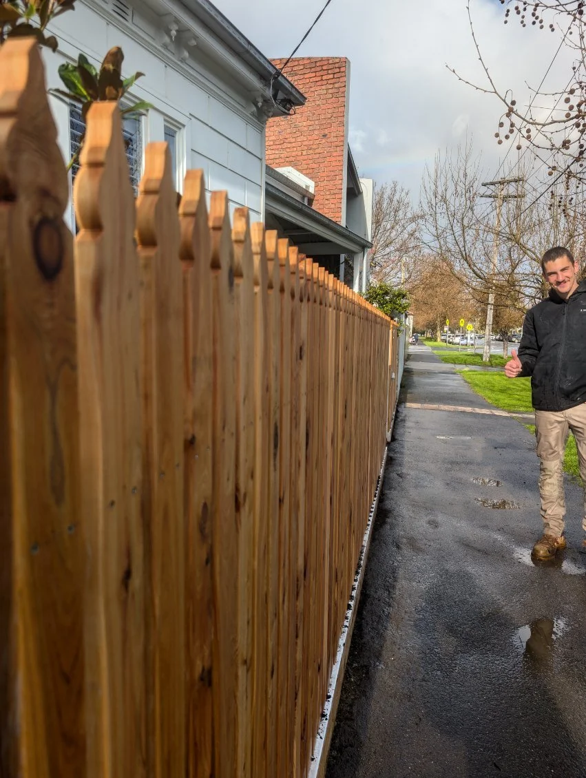 A man in a black jacket and tan pants giving a thumbs-up while walking along a wet sidewalk beside a wooden fence, with houses, trees, and a cloudy sky in the background.