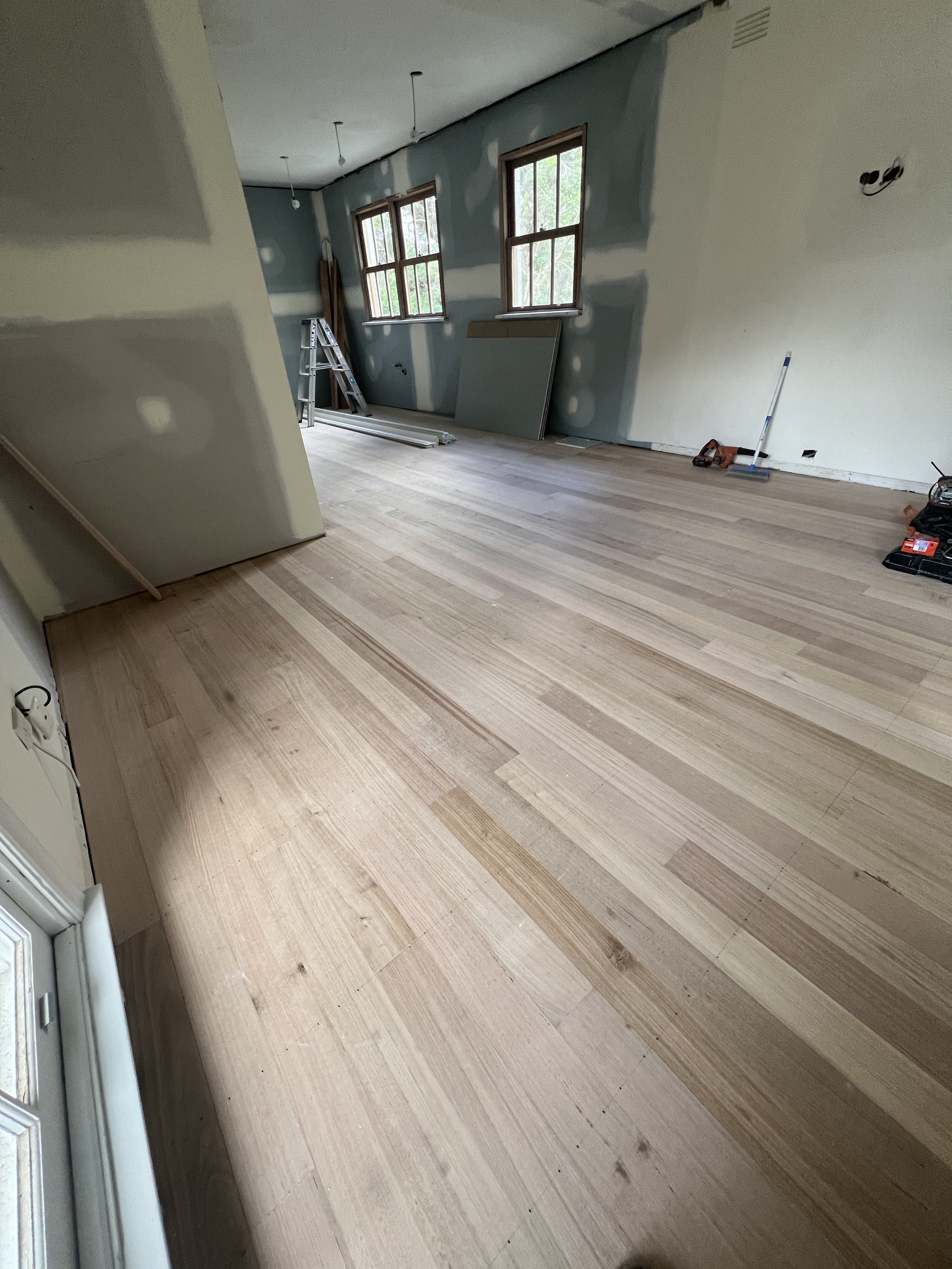 Interior room under renovation with new hardwood flooring, unpainted walls, construction tools, ladder, and windows.