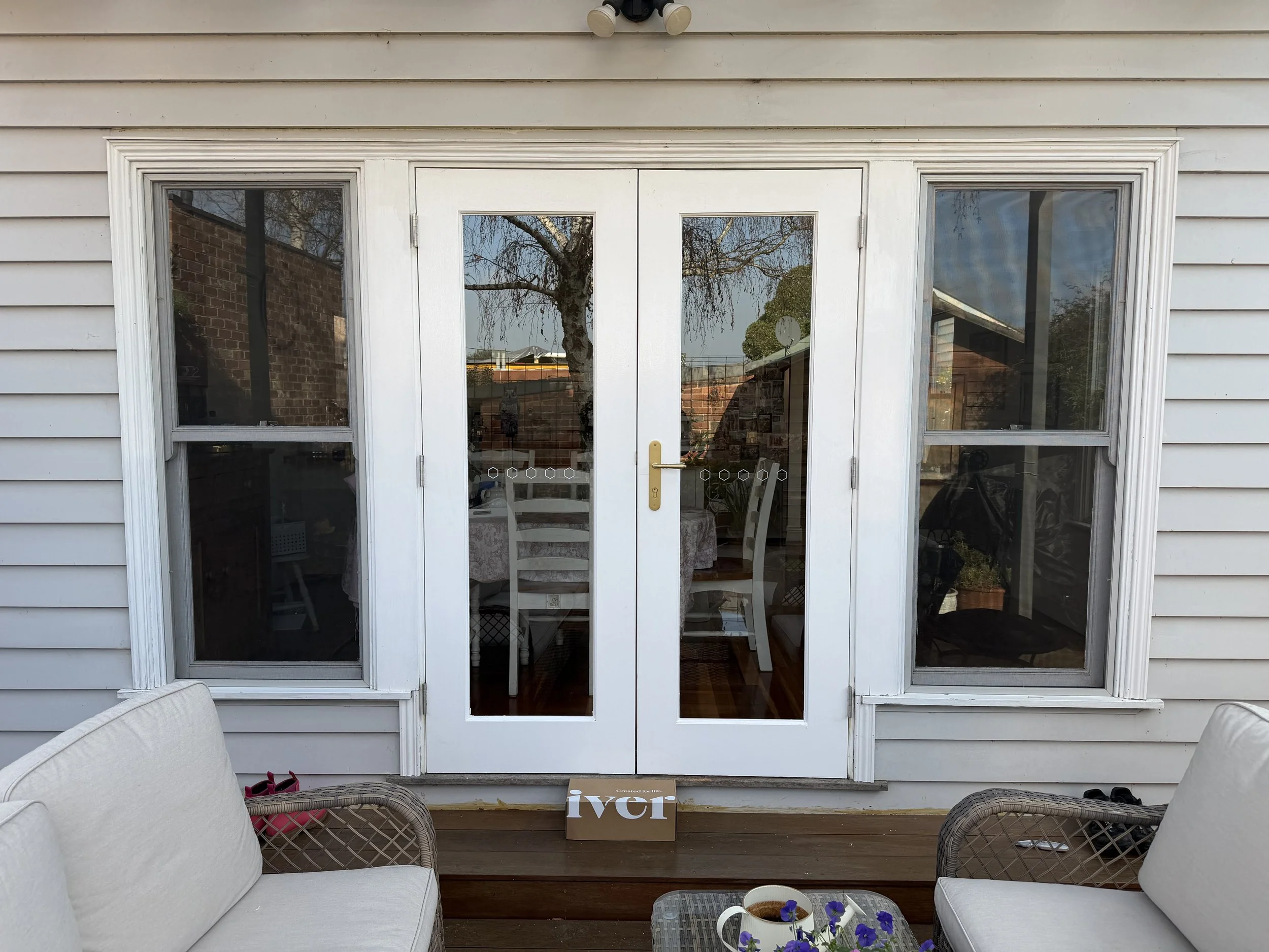 White patio doors with glass panes and a brass handle, framed by white vinyl siding, reflecting a tree and neighboring structures, with patio furniture visible inside.