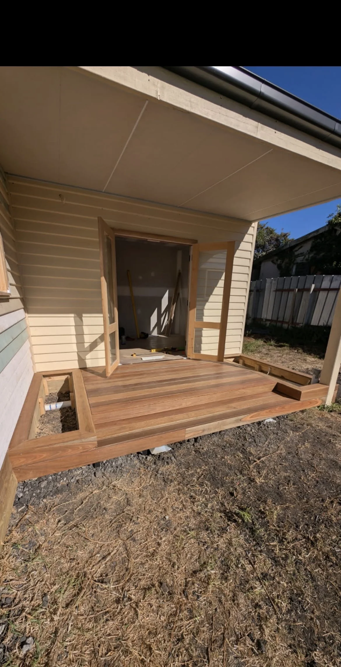 Construction of a wooden deck outside a house with open double doors leading inside. The deck is partially built, with some framing and tools visible inside the house.