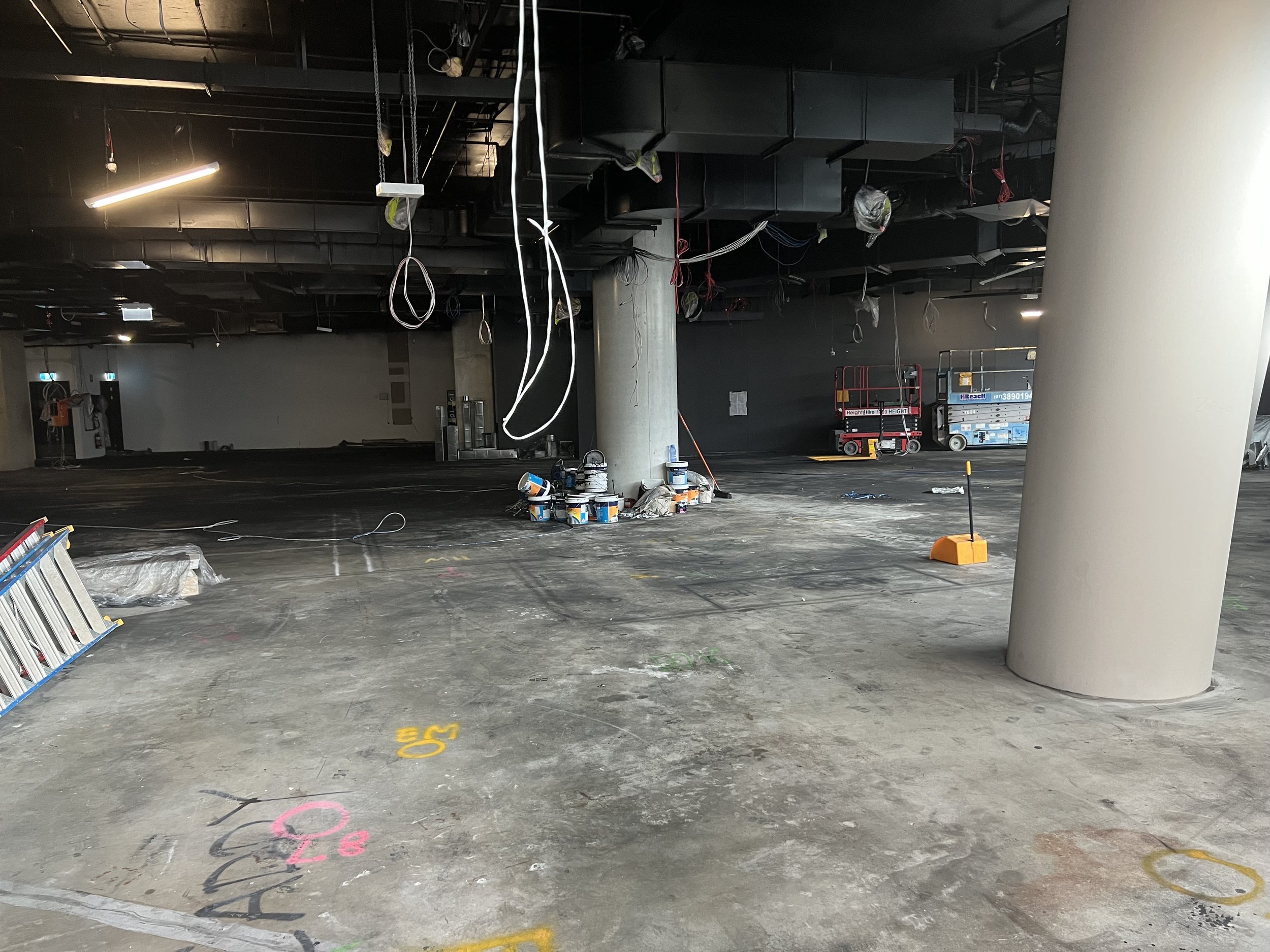 Interior view of a construction site with black ceiling panels, hanging wires, and construction equipment. The floor has colored markings.