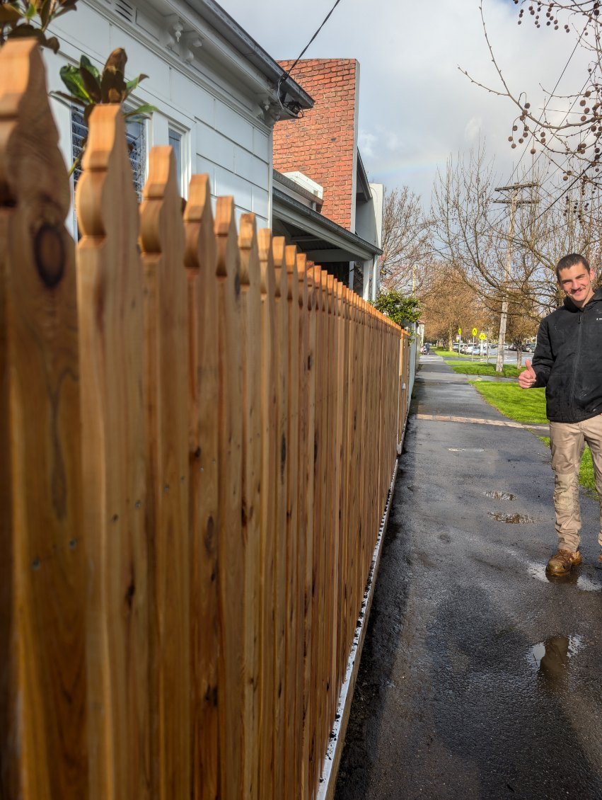 A wooden fence alongside a paved sidewalk on a residential street. A young man is walking on the sidewalk, giving a thumbs-up, with a neighborhood of houses and leafless trees in the background. The sky is partly cloudy with a faint rainbow visible.