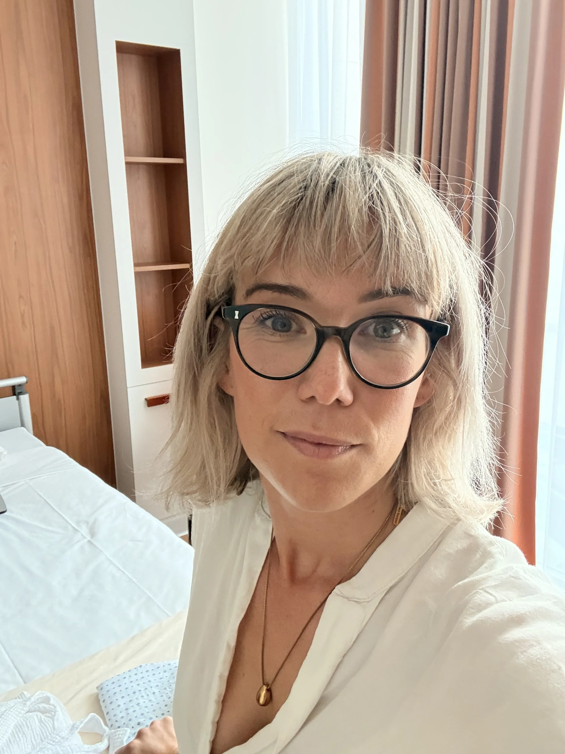 A woman taking a selfie in a bedroom, wearing glasses and a white top, with a head of blonde hair and a necklace, standing near a bed with a white sheet, wooden wardrobe, and curtains in the background.