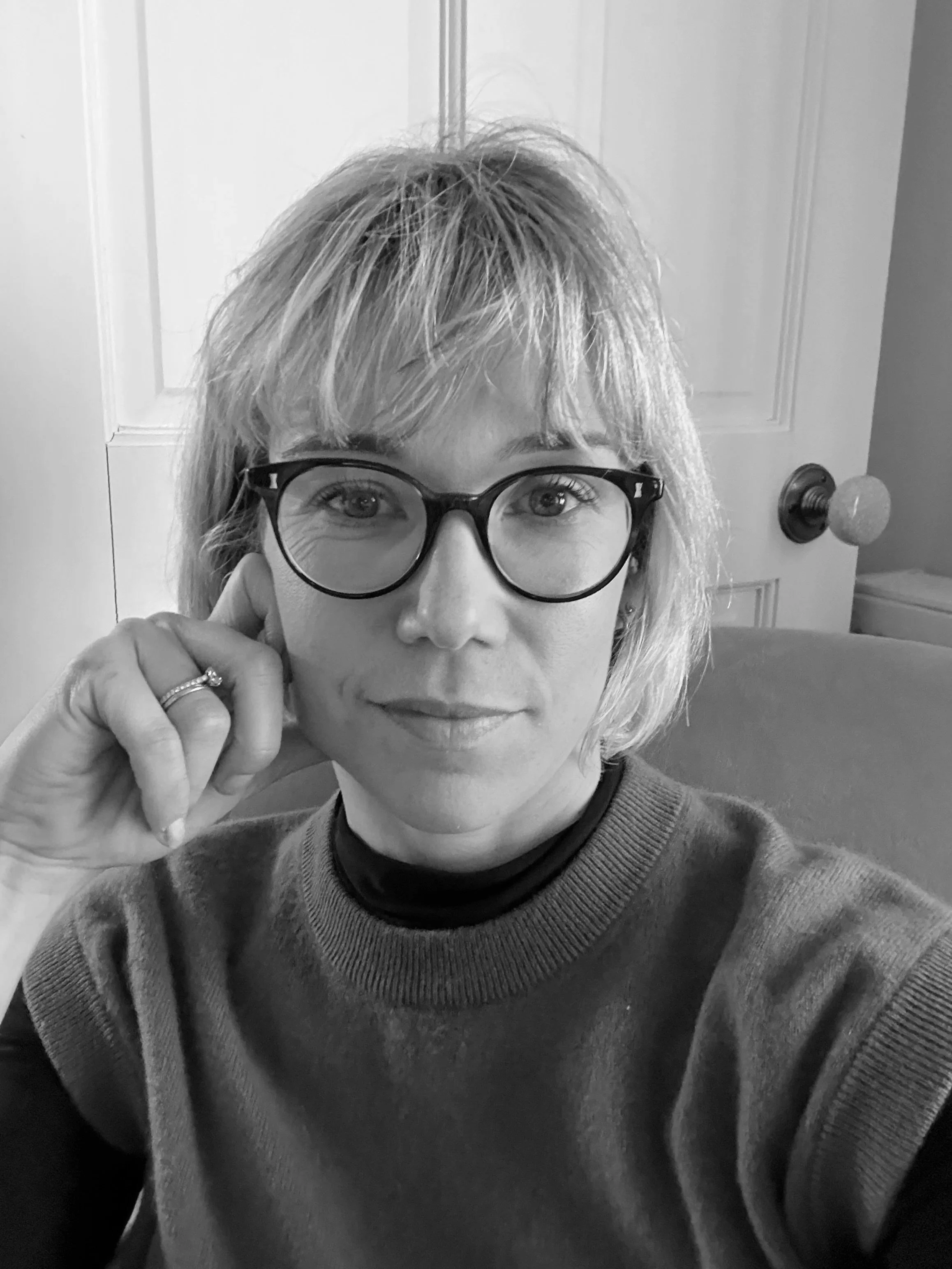 Black and white photo of a woman with blonde hair, wearing glasses, resting her face on her hand, in front of a door.