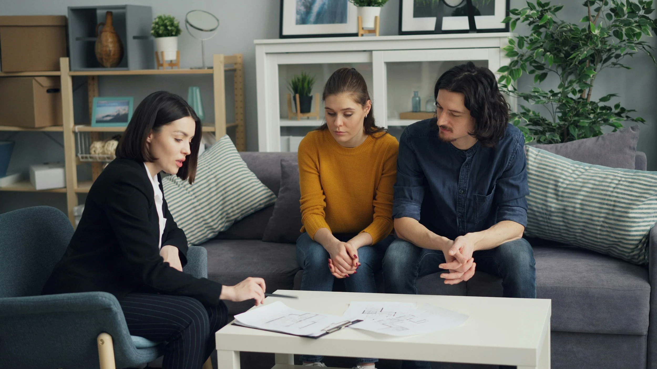 Three adults seated in a living room engaged in a serious discussion, with papers and documents on the table.
