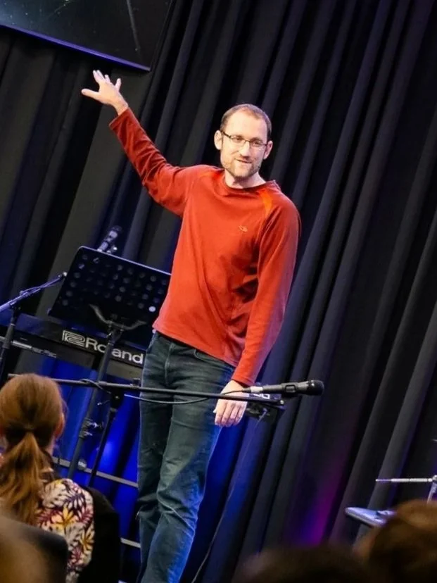A man in a red long-sleeve shirt and glasses standing on a stage with black curtains, gesturing upward with his right hand, with an audience in front of him, and a keyboard on a stand behind him.