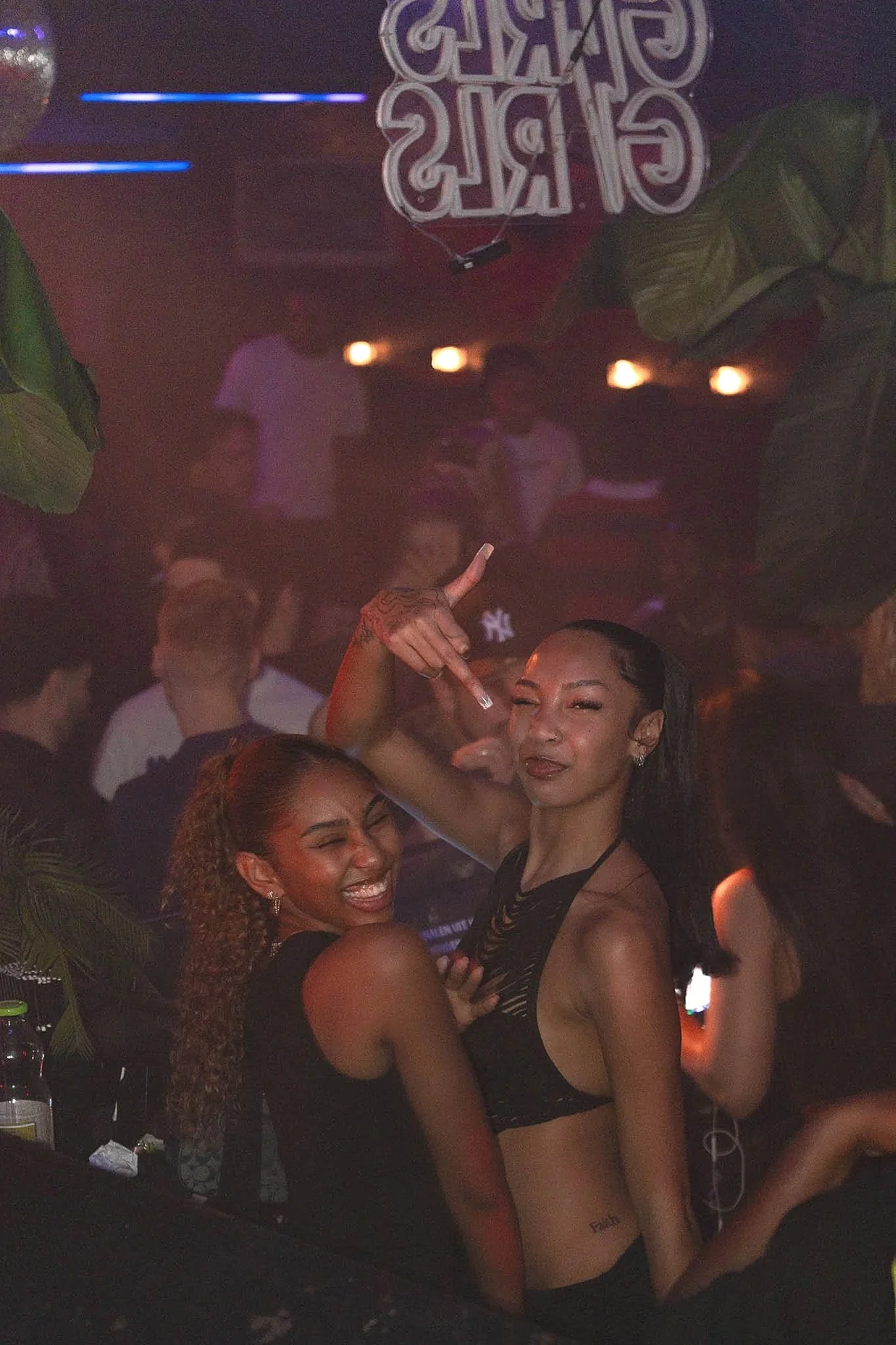 Twee vrouwen in een nachtclub of bar, één glimlachend en de ander met een speelse blik, omringd door mensen en kleurrijke lichten.