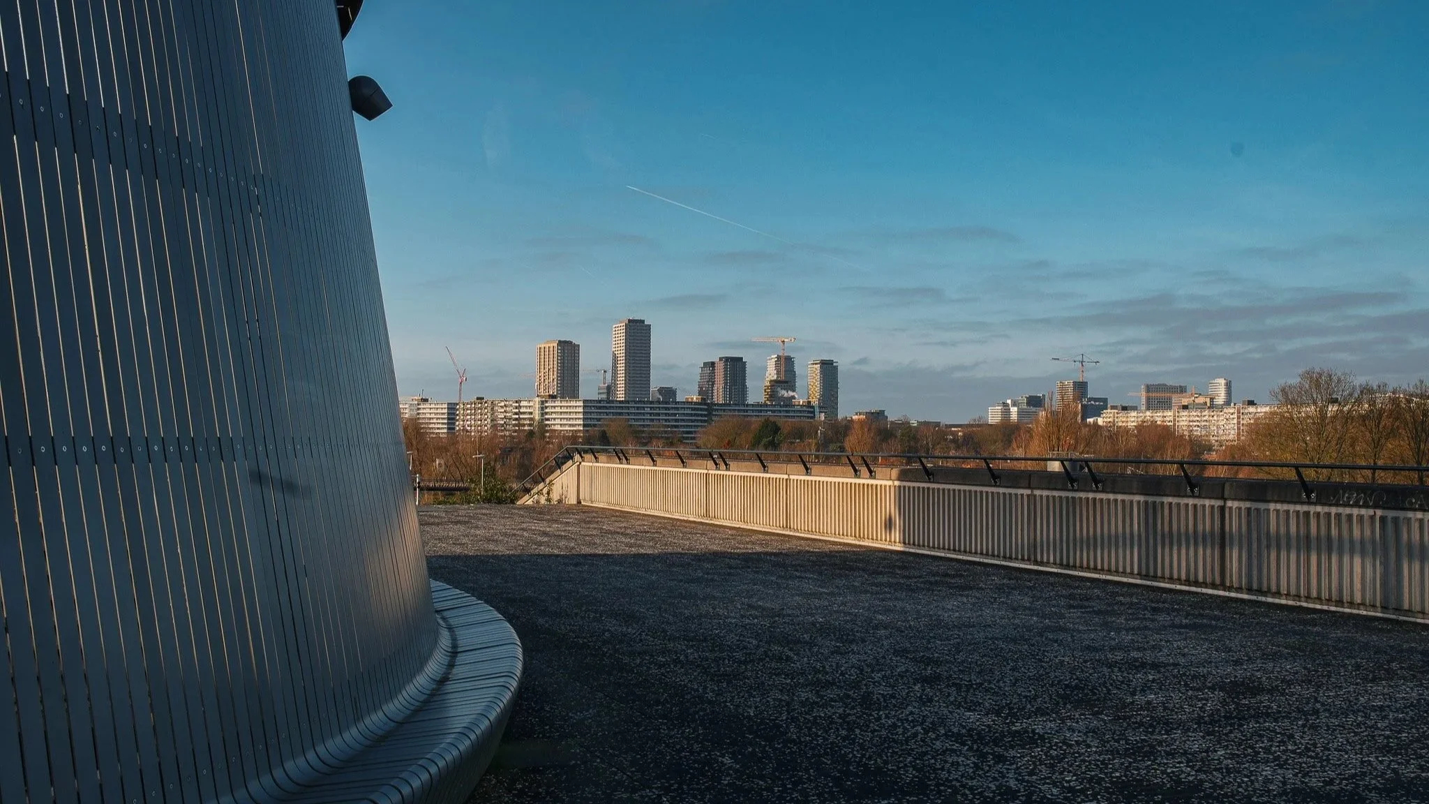 Uitzicht van een modern stadsgebouw met metalen gevel en een balkon, met een skyline van hoge gebouwen en een blauwe hemel op de achtergrond.
