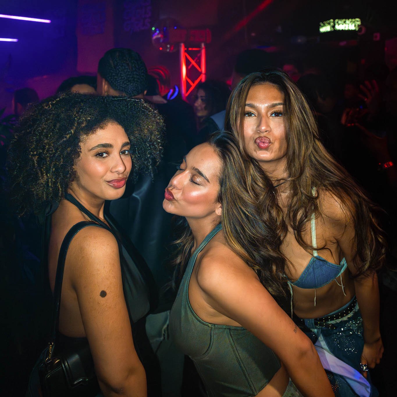 Drei vrouwen in een nachtclub die samen poseren, met kleurrijk neonlicht en andere mensen op de achtergrond.