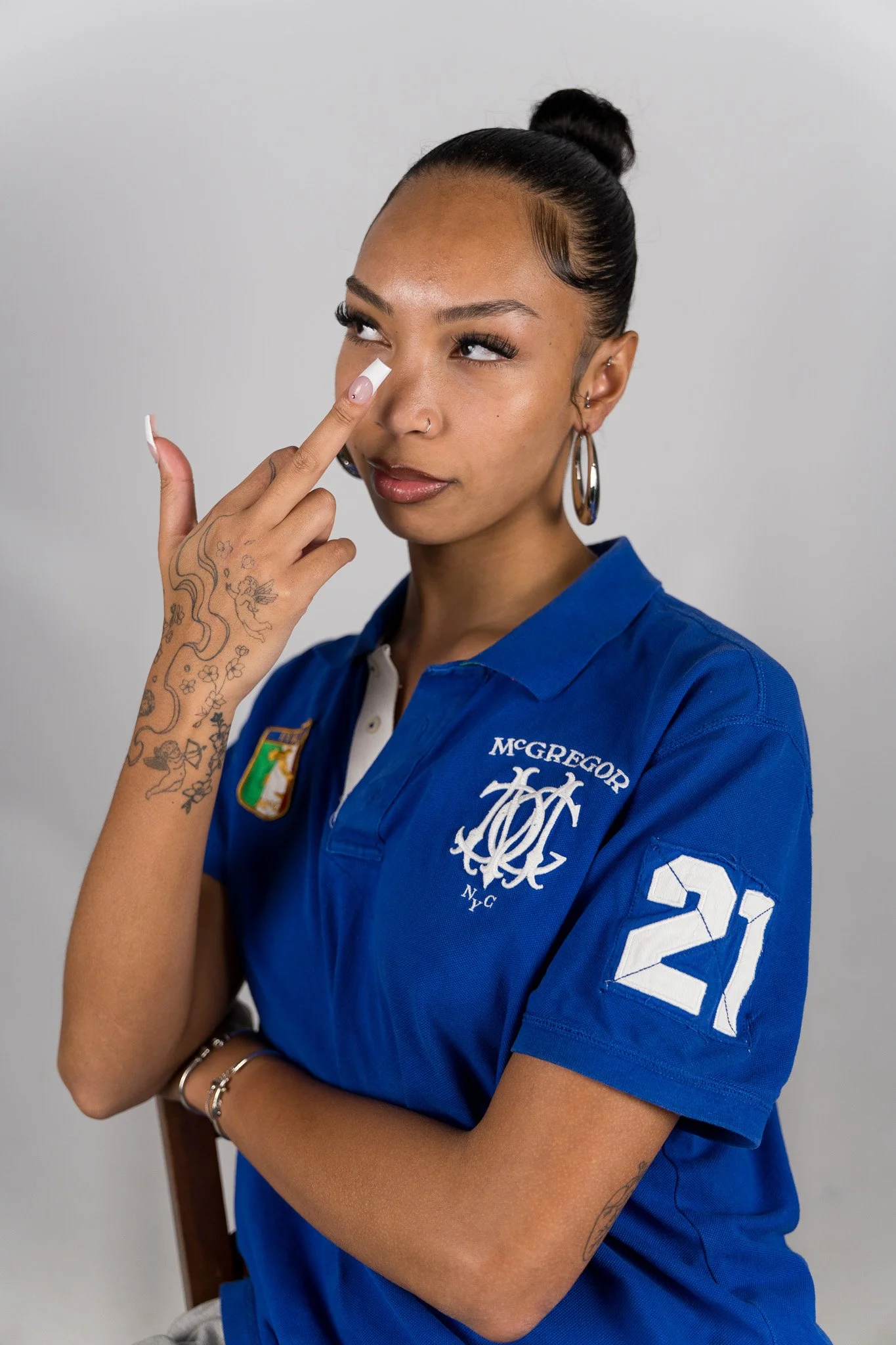Vrouw in armkleding van de New York Yankees met tatoeages op haar arm, die haar vinger op haar neus plaatst, gekleed in een blauwe sportshirt met logo en nummer 21.