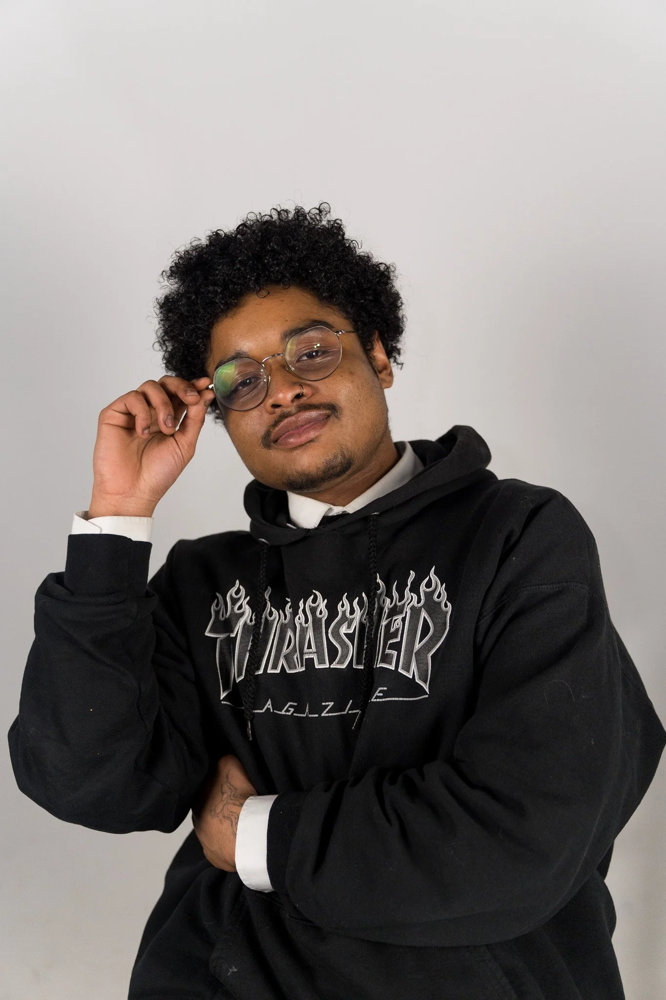 Portret van een jonge man met krullend zwart haar, bruin huid, ronde glazen, een nepneus en een tatoeage op zijn hand, gekleed in een zwarte hoodie met een grafisch ontwerp, tegen een witte achtergrond.