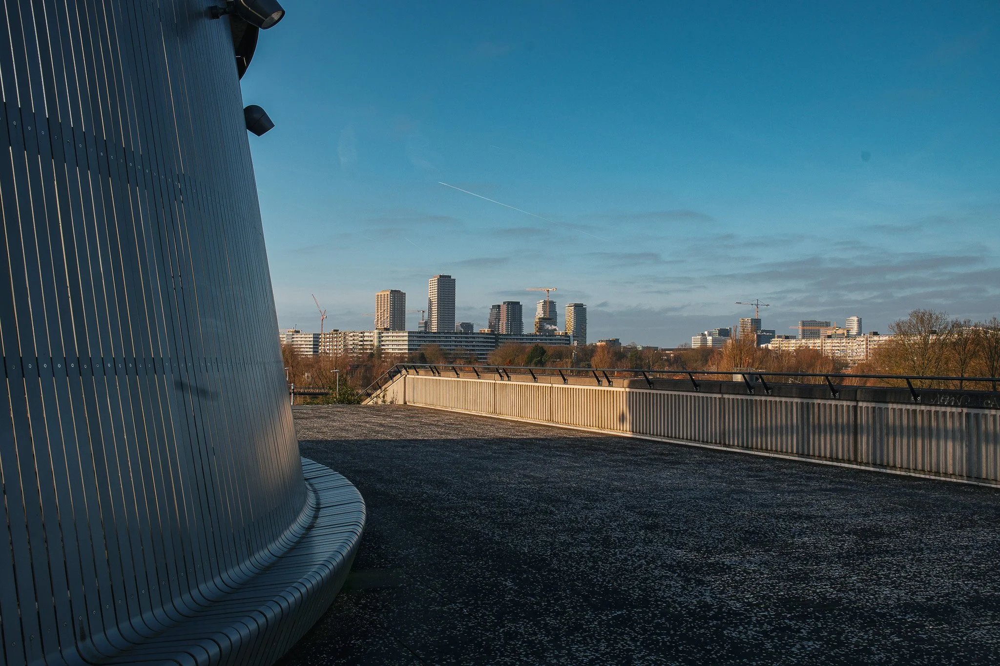Uitzicht van een modern stadsgebouw met metalen gevel en een balkon, met een skyline van hoge gebouwen en een blauwe hemel op de achtergrond.