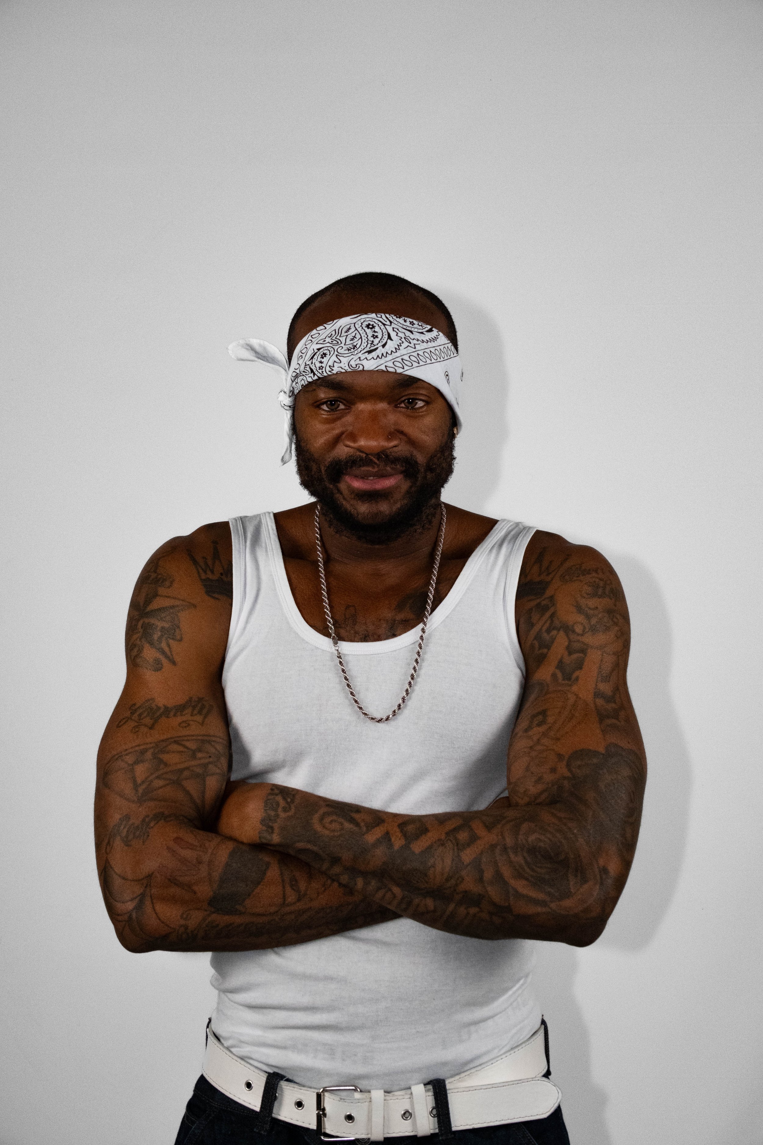 Een man met getatoeëerde armen, een witte tanktop en een witte gordel, met een witte bandana op zijn hoofd, staat voor een witte muur. Hij kijkt recht in de camera met een zelfverzekerde uitdrukking.