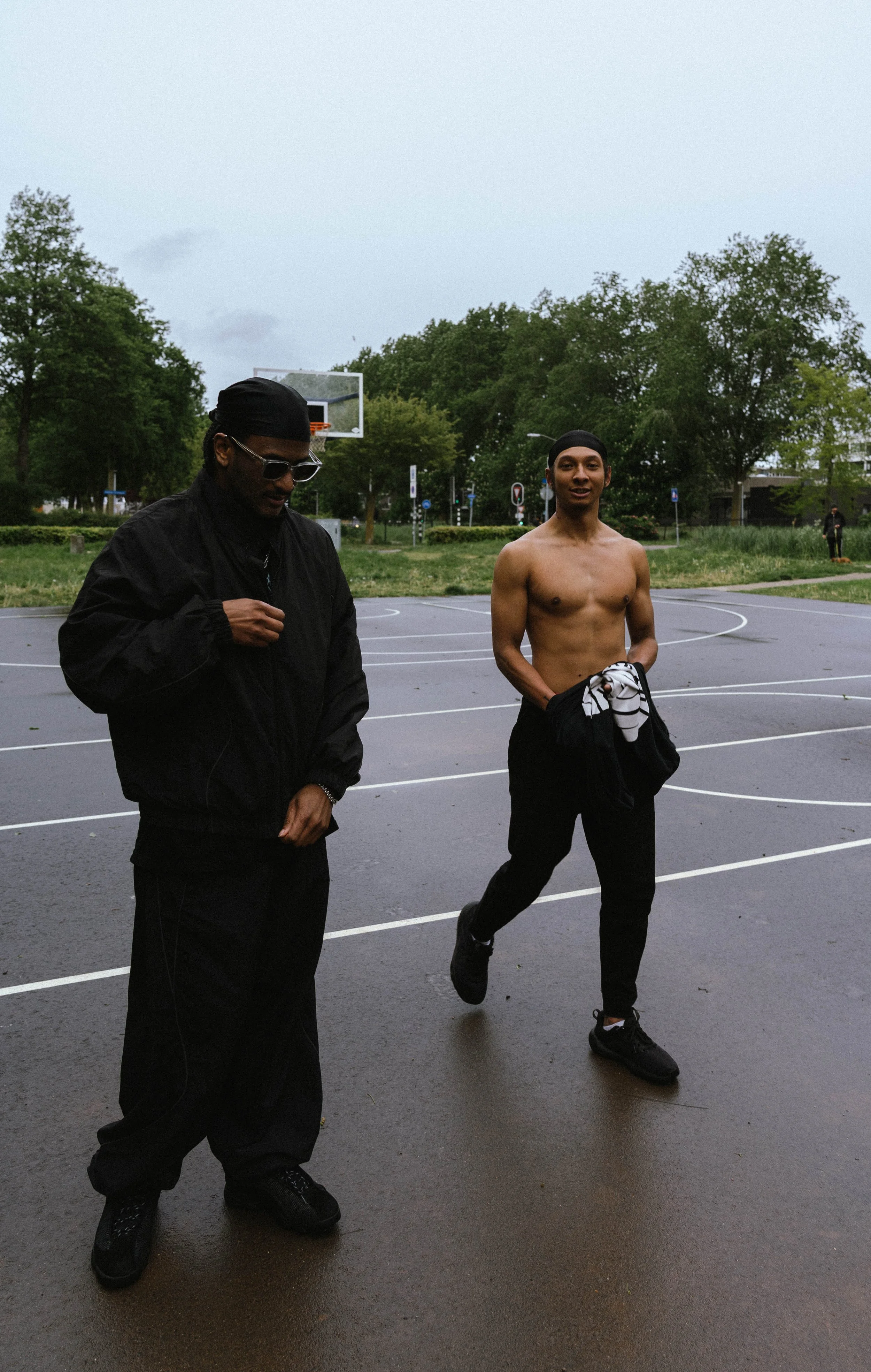 Twee jonge mannen, één shirtloos met een zwart hoofddeksel en een ander gekleed in een zwarte jas en zonnebril, wandelen op een basketballcourt met groene bomen en sportborden op de achtergrond.