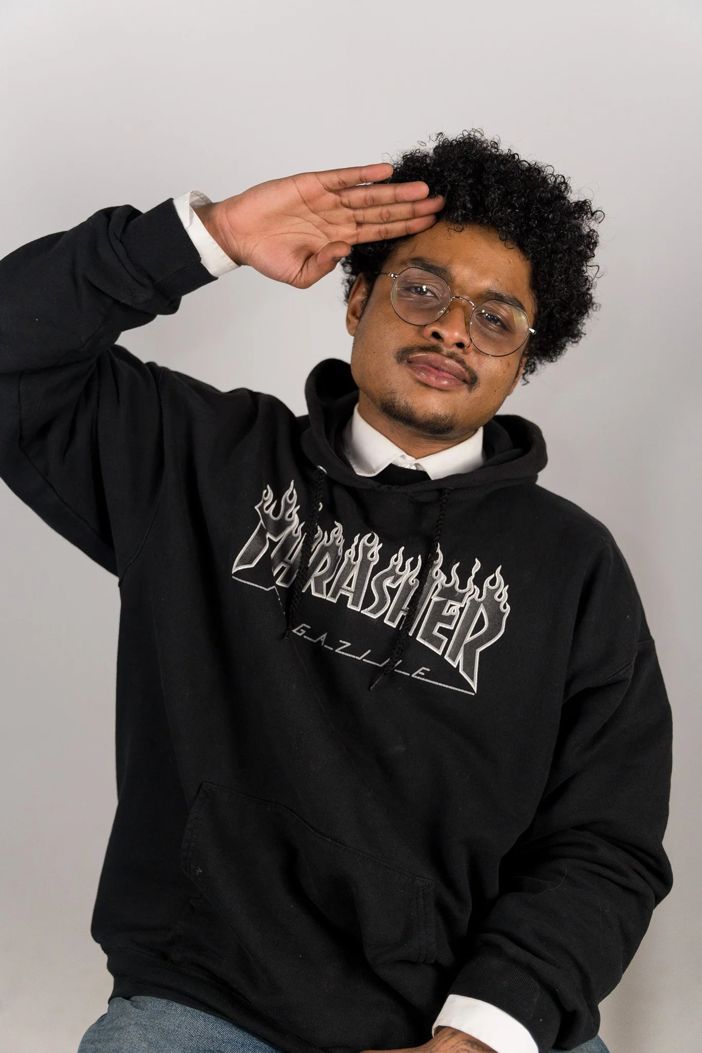 Een man met krullend haar, bril en een lichte huid, draagt een zwarte hoodie met een grafisch ontwerp en poseert met een saluut gebaar tegen een lichte achtergrond.