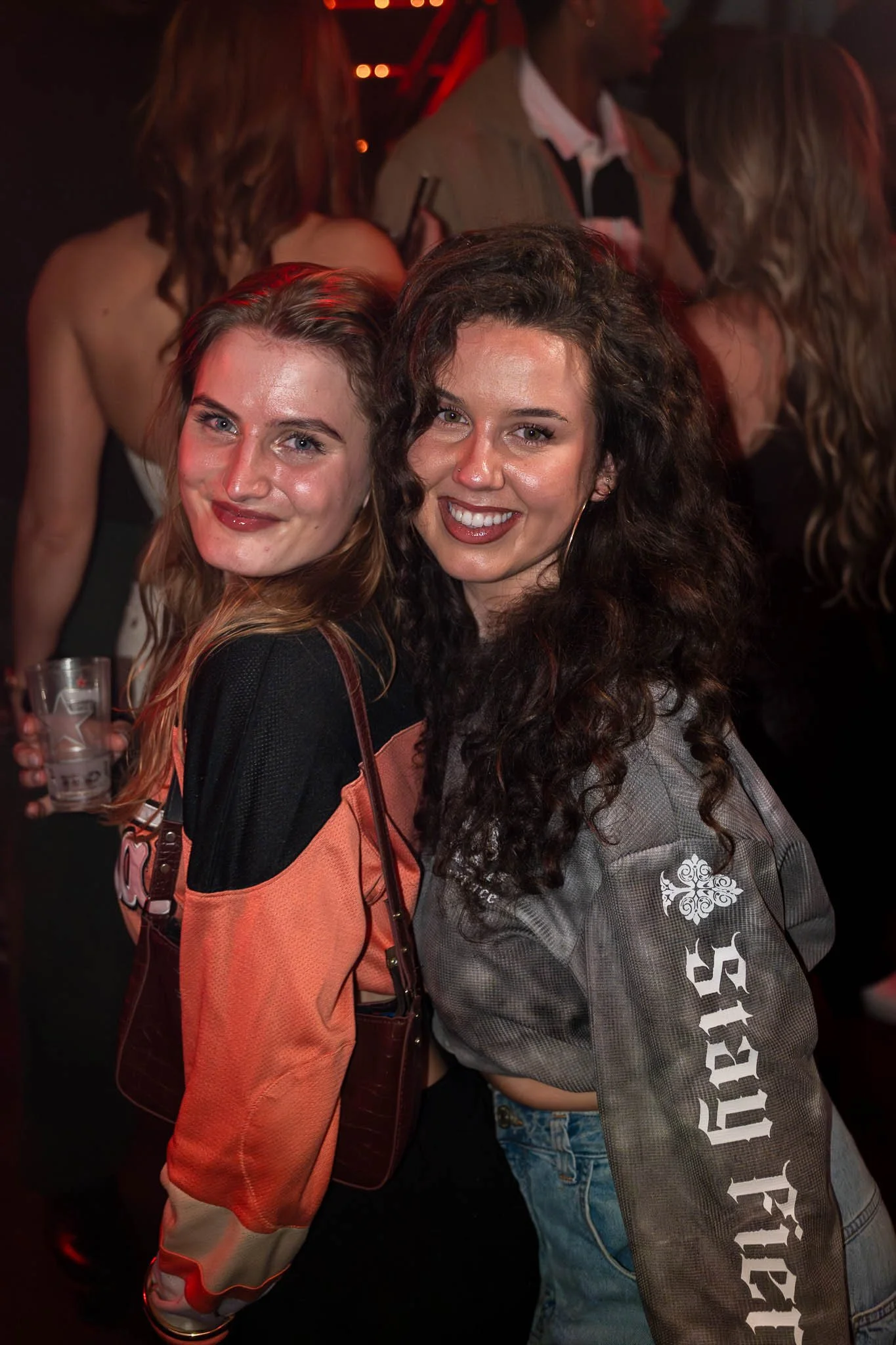 Twee jonge vrouwen poseren in een donkere sfeer met rode en blauwe lichten, lachend en genietend op een feestje of club.