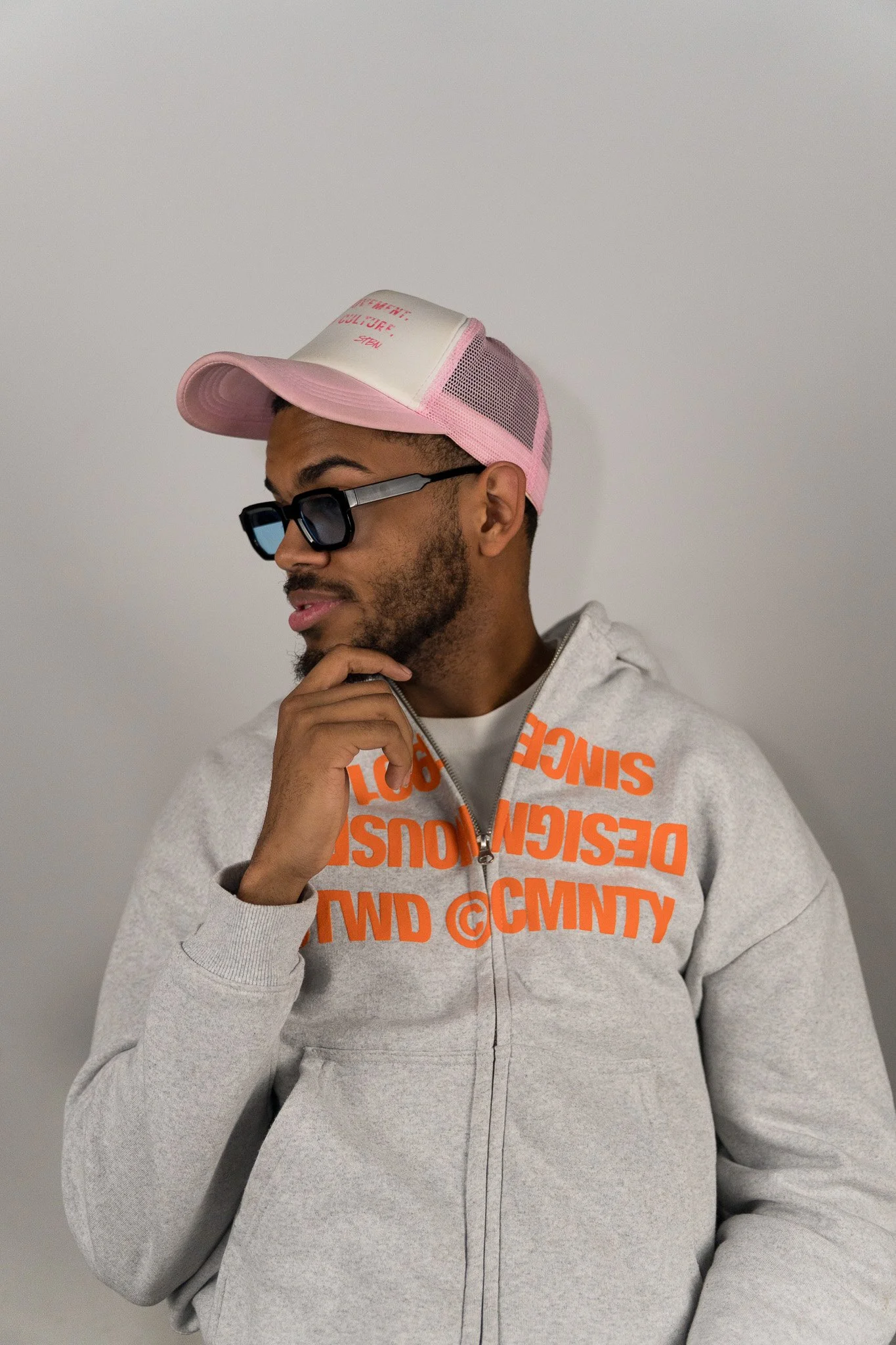 Man met zwart haar, een korte baard en een mustache, draagt een lichtgrijze hoodie met oranje tekst, een roze en witte baseballpet en zwarte zonnebril. Hij staat tegen een effen lichtgrijze achtergrond.