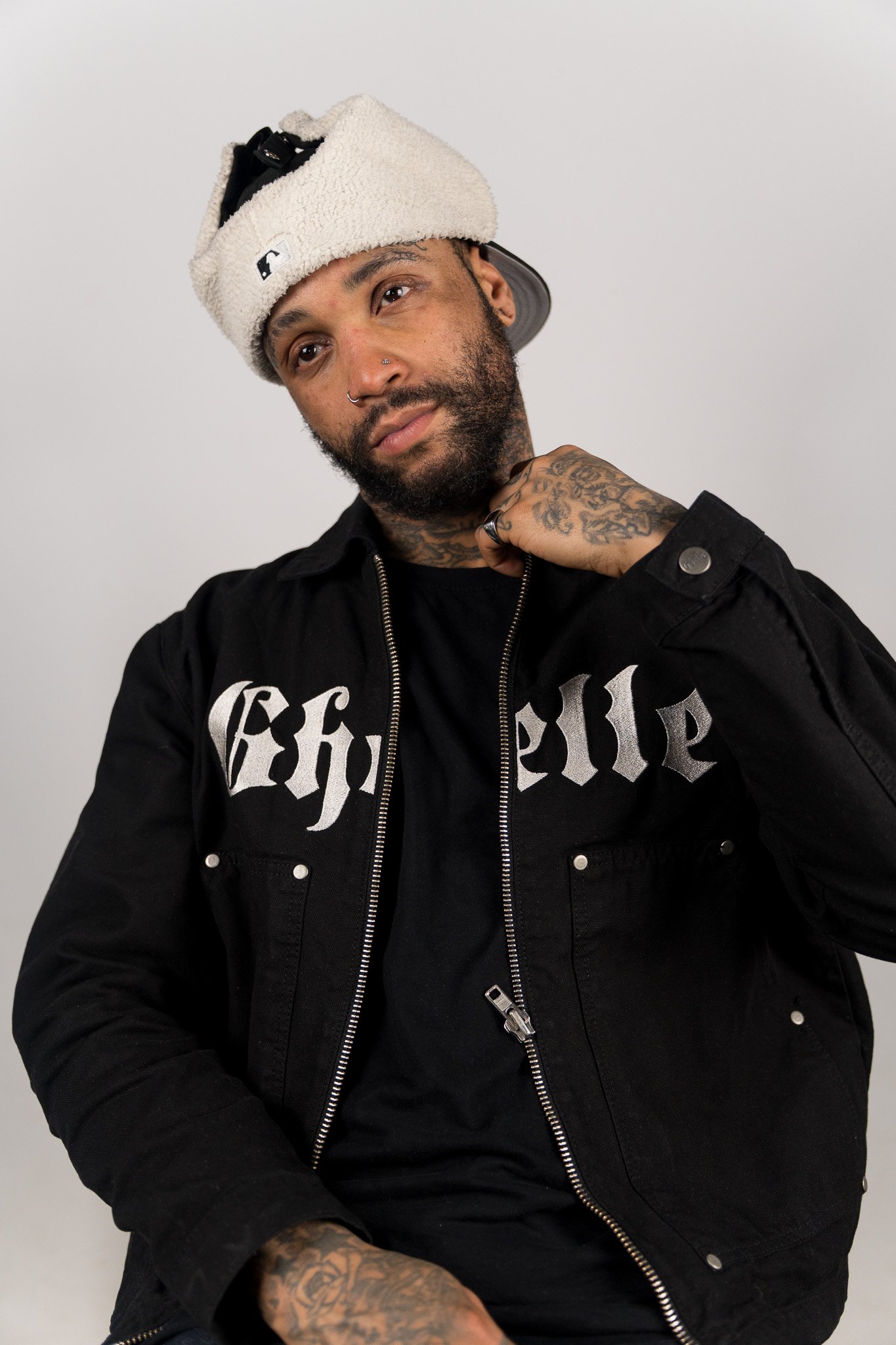 Een man met een pet die ondersteboven op zijn hoofd ligt, een black jacket met grote witte letters, en een tattoo op zijn arm, zit tegen een witte achtergrond.