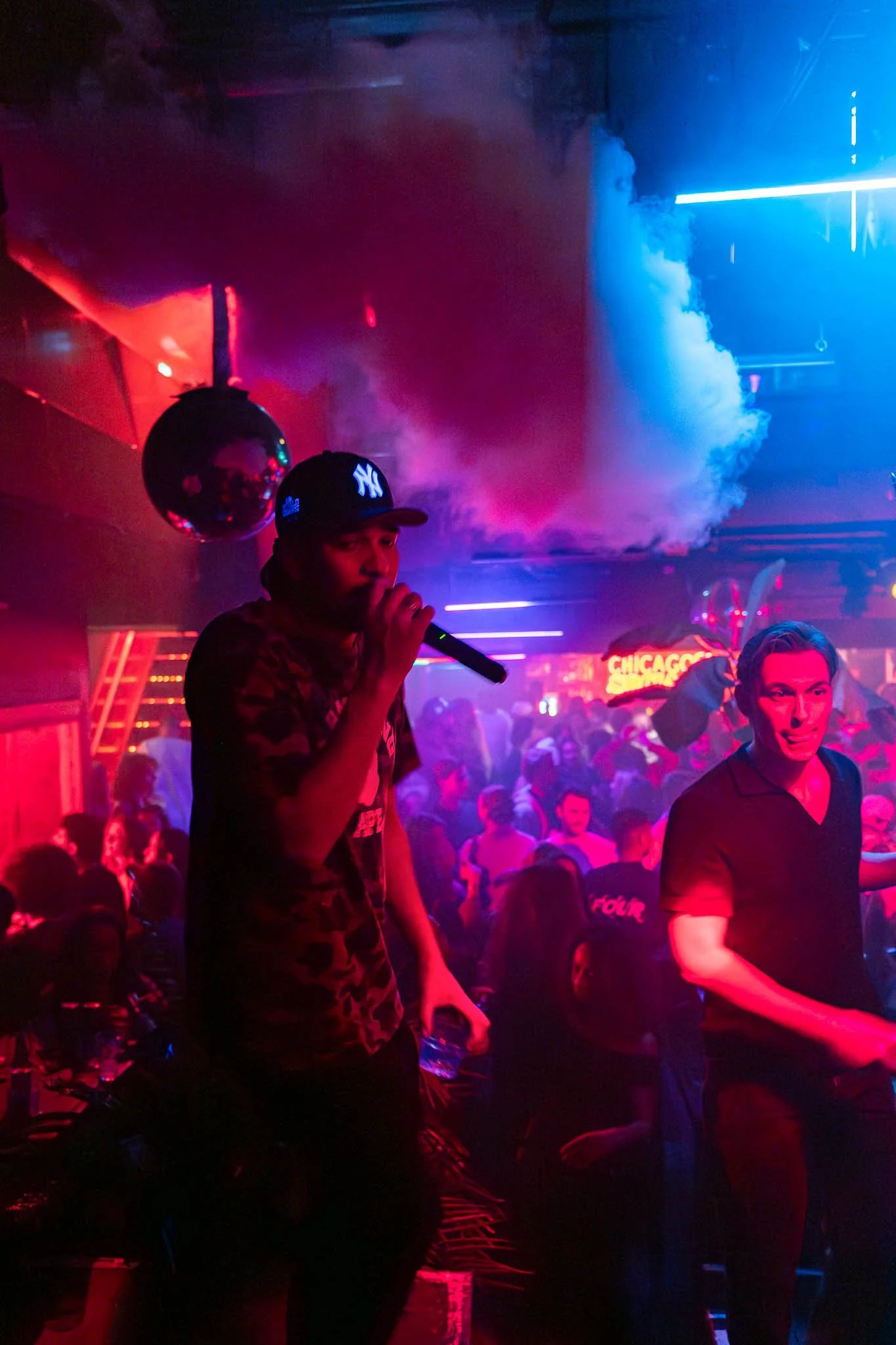 Een drukke clubscene met mensen die dansen en plezier hebben, onder kleurrijke neonlichten en rookmachines.