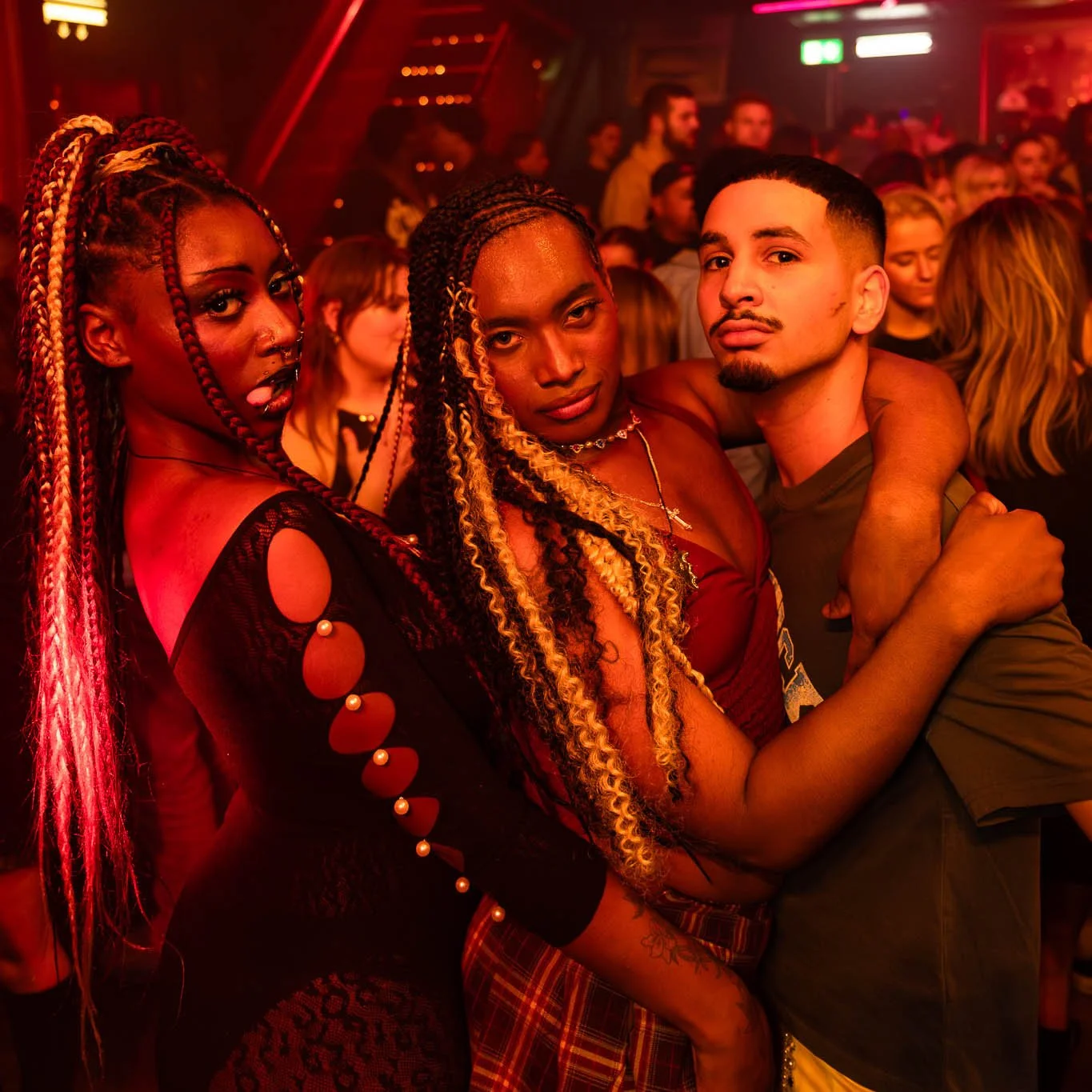Drie mensen dansen en poseren in een donkere, rood verlichte omgeving, mogelijk een nightclub of feest. De sfeer is levendig en feestelijk.