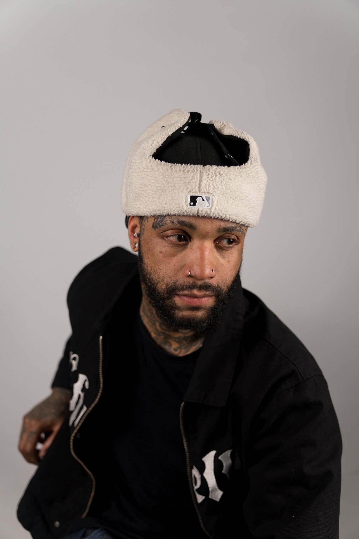 Portret van een man met een baard en tattoos, die een zwart hoodie en een beige teddy pet draagt, met een sprekend gezicht en een glimlach.