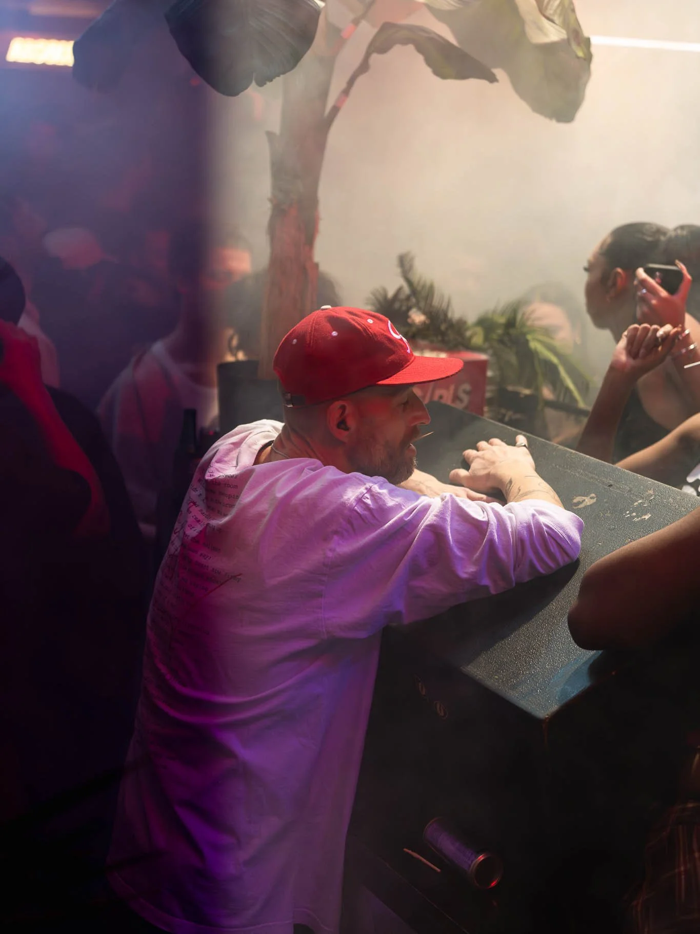 Man met een rode baseballpet en een witte hoodie leunt op een tafel in een club, terwijl een vrouw met een telefoon voor haar gezicht in de achtergrond staat.