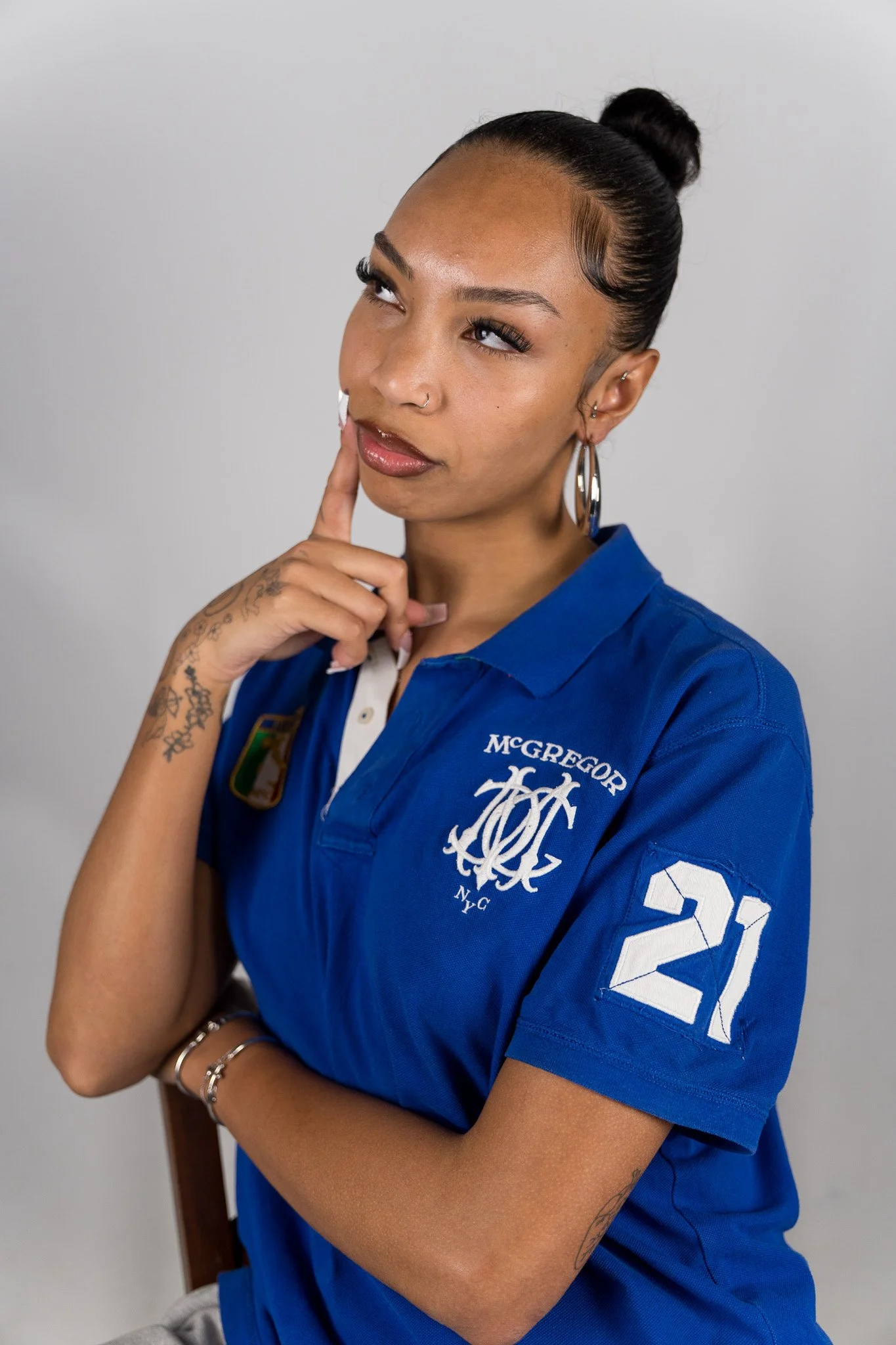 Een vrouw met een hoge paardenstaart, vleugje make-up en meerdere oorbellen, draagt een blauwe sportshirt met het logo van McGregor NY en het nummer 21 op de mouw, terwijl ze nadenkend met haar vinger op haar lippen ligt.