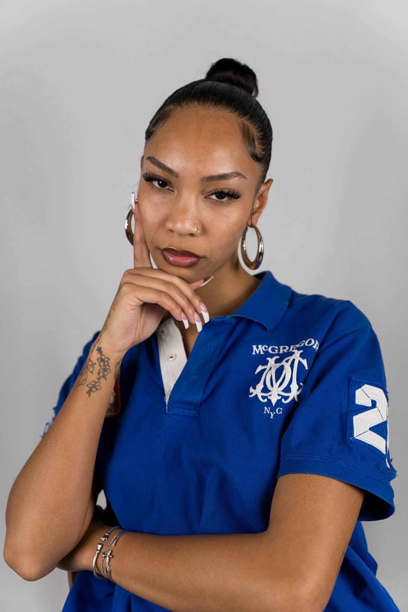 Portret van een vrouw met donkere haren in een strakke knot, grote oorbellen, een neusring, en meerdere sieraden, gekleed in een blauwe rugbyjas met logo's, poseert met haar hand onder haar kin tegen een grijze achtergrond.