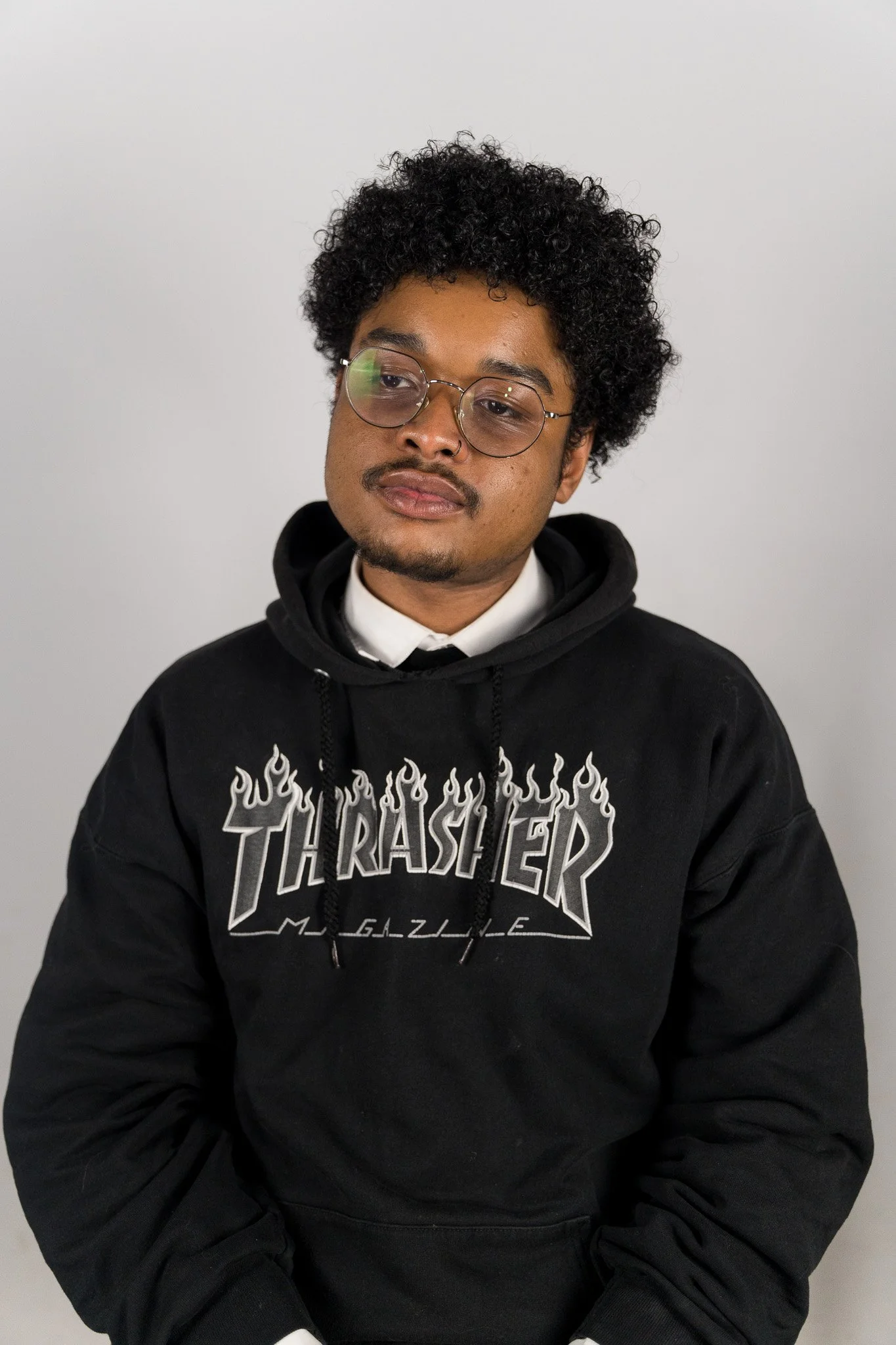 Portretfoto van een jonge man met kort, krullend haar en bril, gekleed in een black hoodie met een Thrasher logo, zittend voor een witte achtergrond.