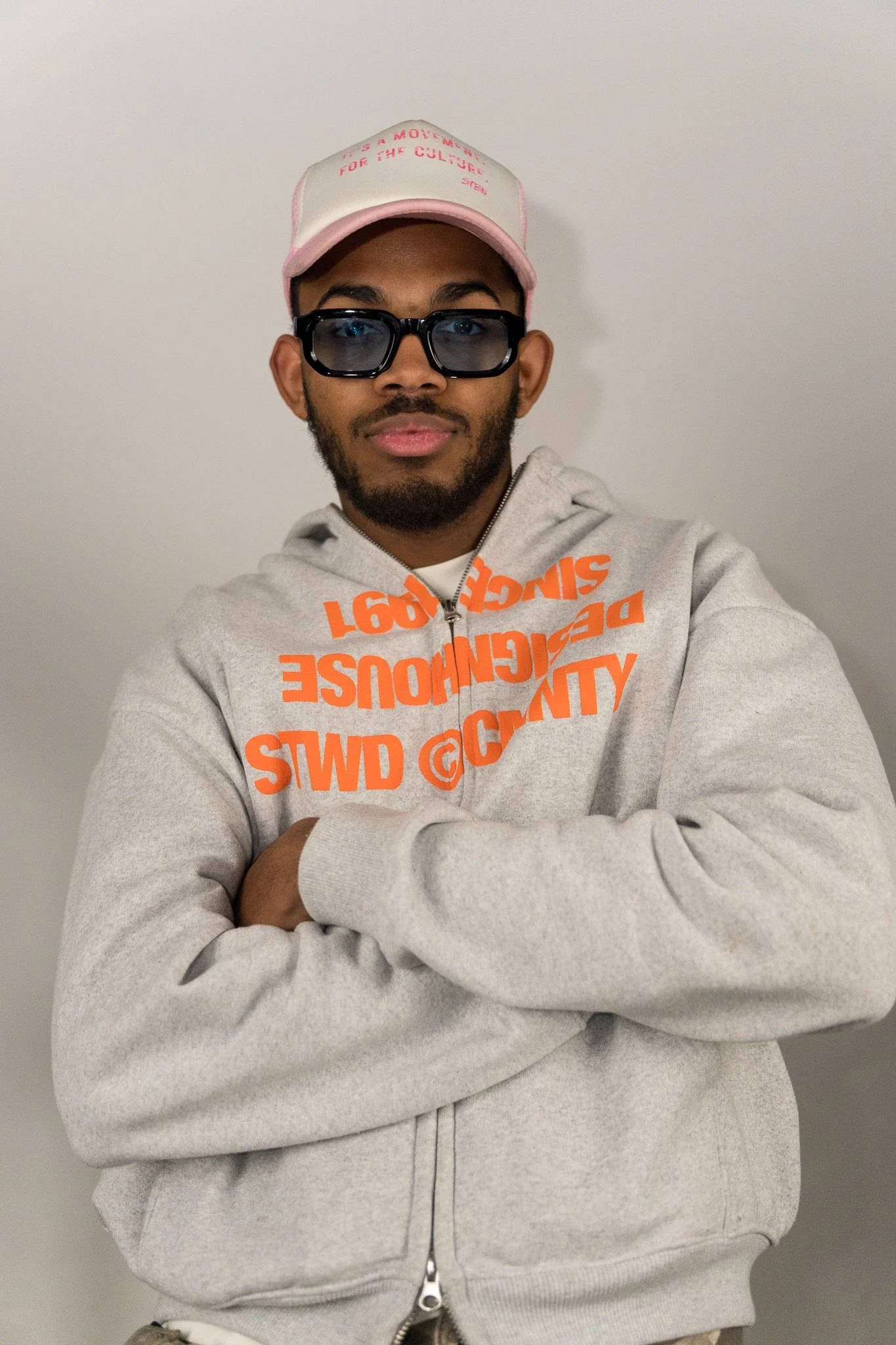 Man draagt een grijze hoodie met oranje tekst, een roze en witte pet en zwarte brillen, staat voor een witte muur.