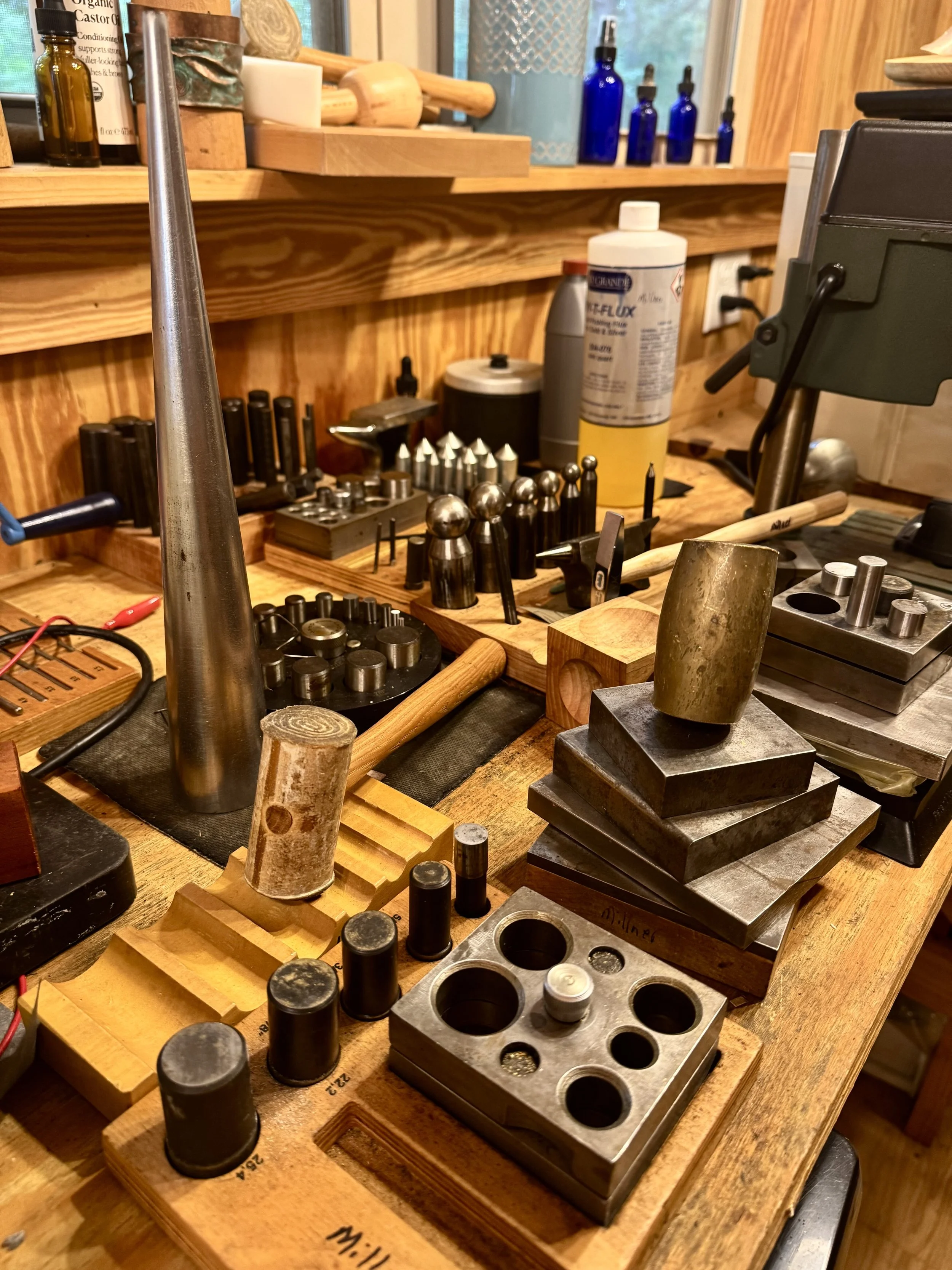 A cluttered workbench with various metal and wooden tools, weights, measuring devices, and bottles, situated in a workshop with wooden walls.