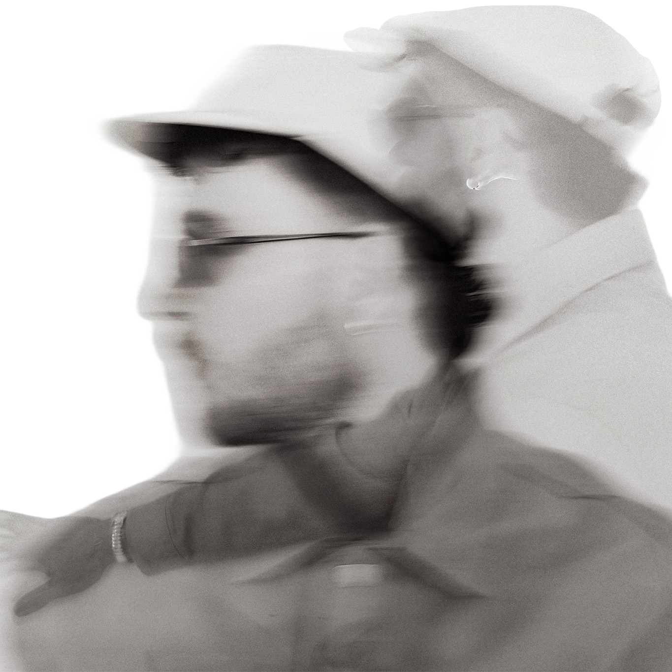 Blurred black and white photo of a person wearing glasses, a hat, and a necklace, with their head turned to the side.